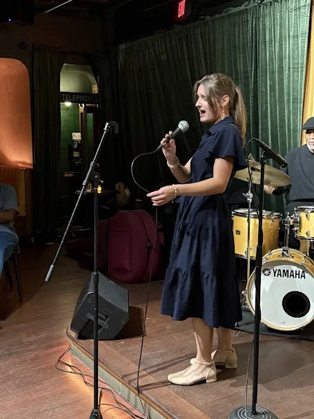 Young woman singing into a microphone on stage with drums and green curtains in the background. Carolyn Kashmanian, Jazz, Live music, Charlotte, North Carolina, bebop, singers, vocalists, eighty eights, Petra's