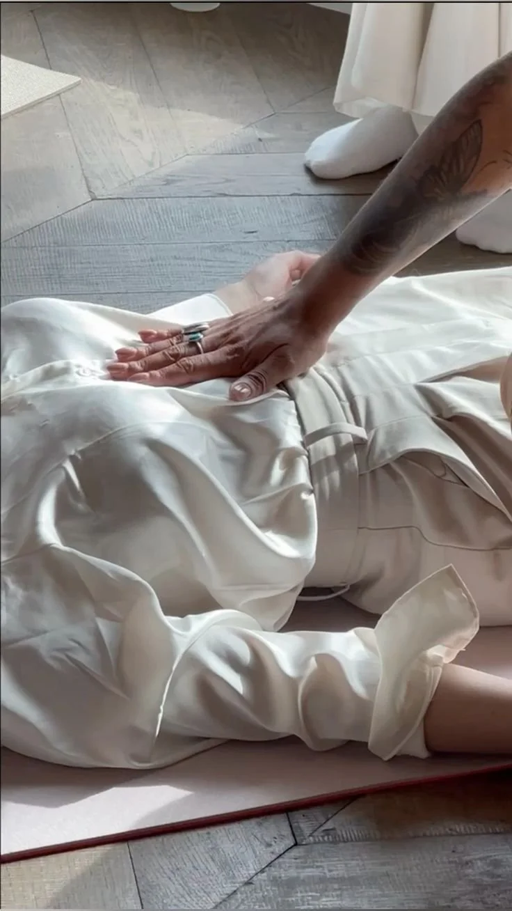 A person wearing white satin pajama pants lying on the floor, with another person placing their hand on their stomach.