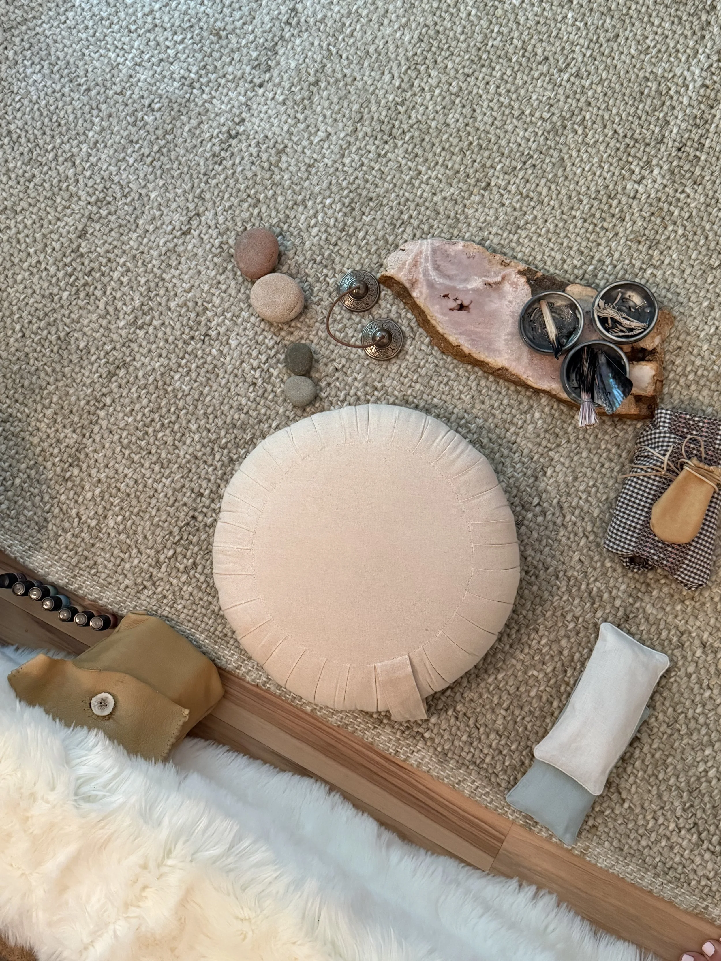 A collection of decorative items on a woven rug, including a round beige cushion, a small pouch, a piece of pink and brown mineral, three tealight candles in metallic holders, and assorted stones.