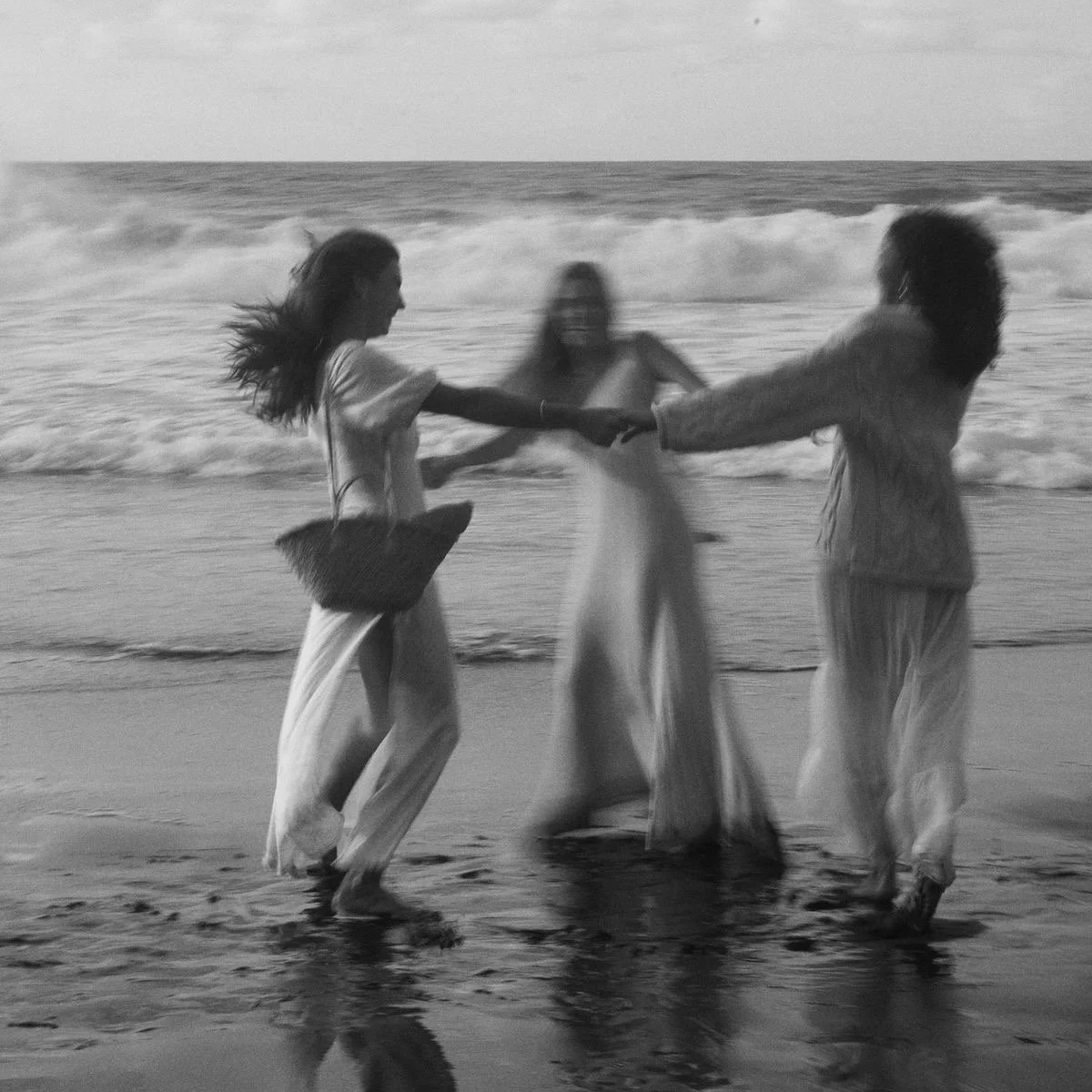 Three women on the beach, holding hands in a circle, with waves in the background, in black and white.