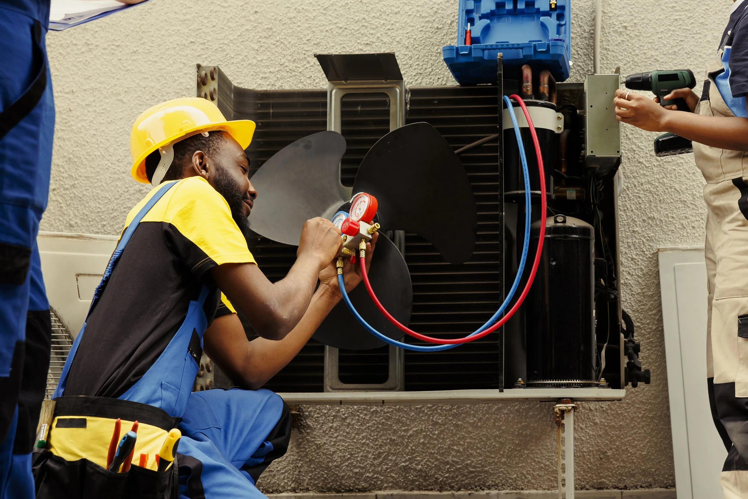 Technician in yellow hard hat and blue overalls repairing an air conditioning unit with gauges, while another technician uses a screwdriver nearby.