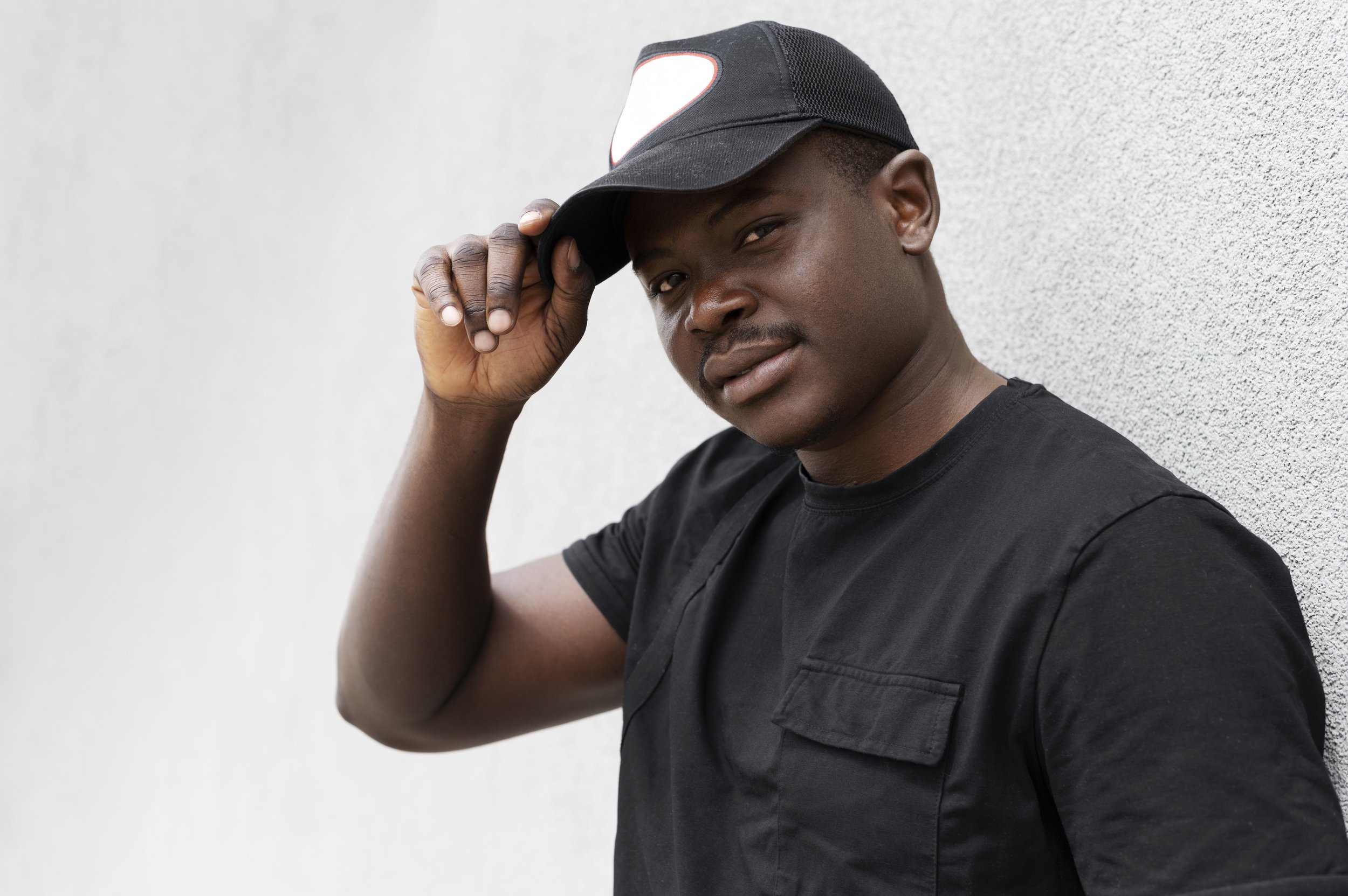 A young African American man in a black t-shirt and cap, standing against a light gray wall, holding the brim of his cap with one hand, looking at the camera.