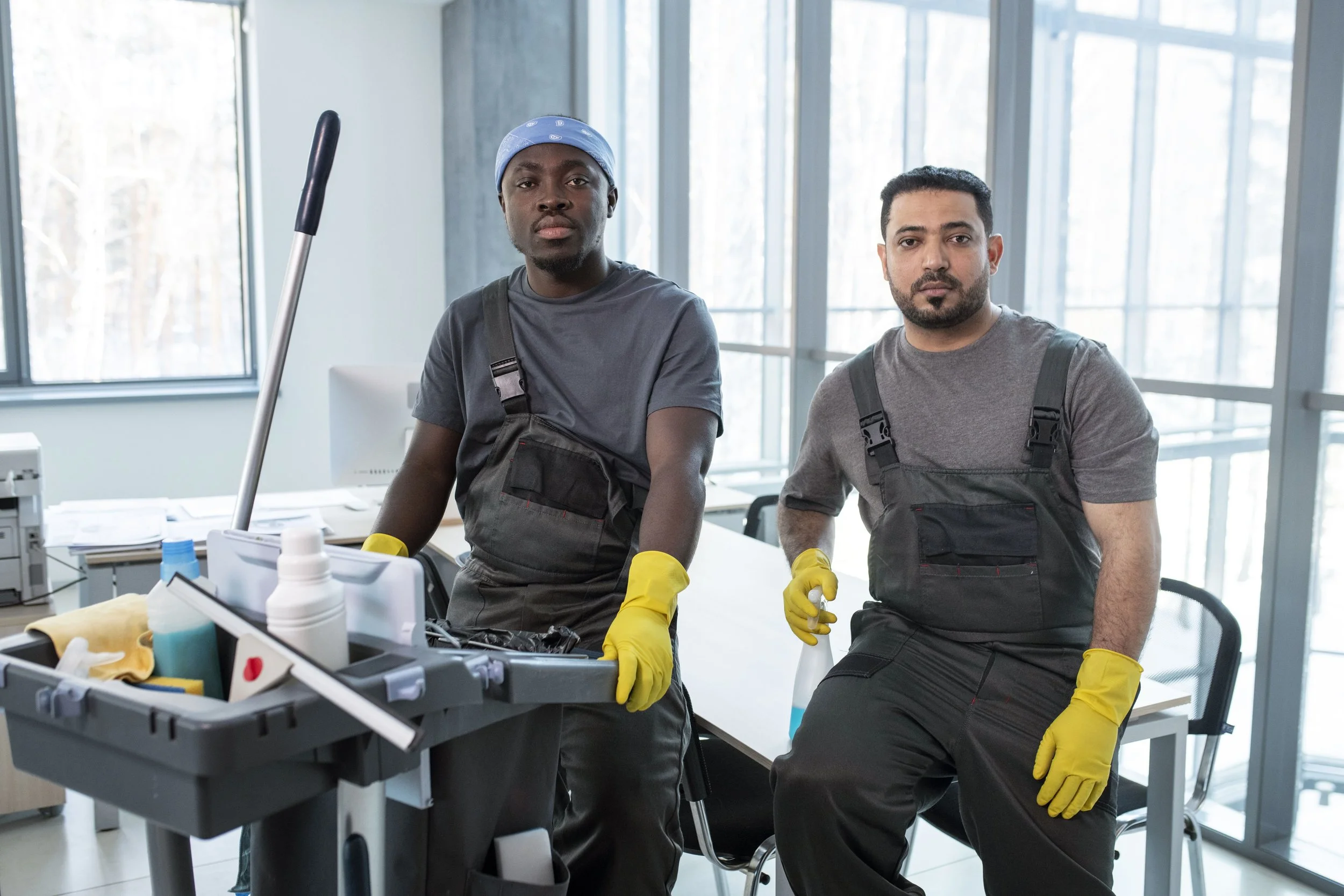 Two male janitors wearing yellow gloves and dark work overalls in a bright modern office cleaning.