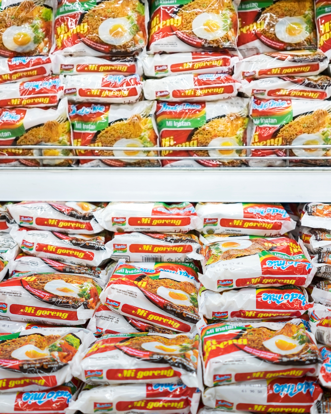 Multiple packets of instant fried noodles stacked on supermarket shelves.