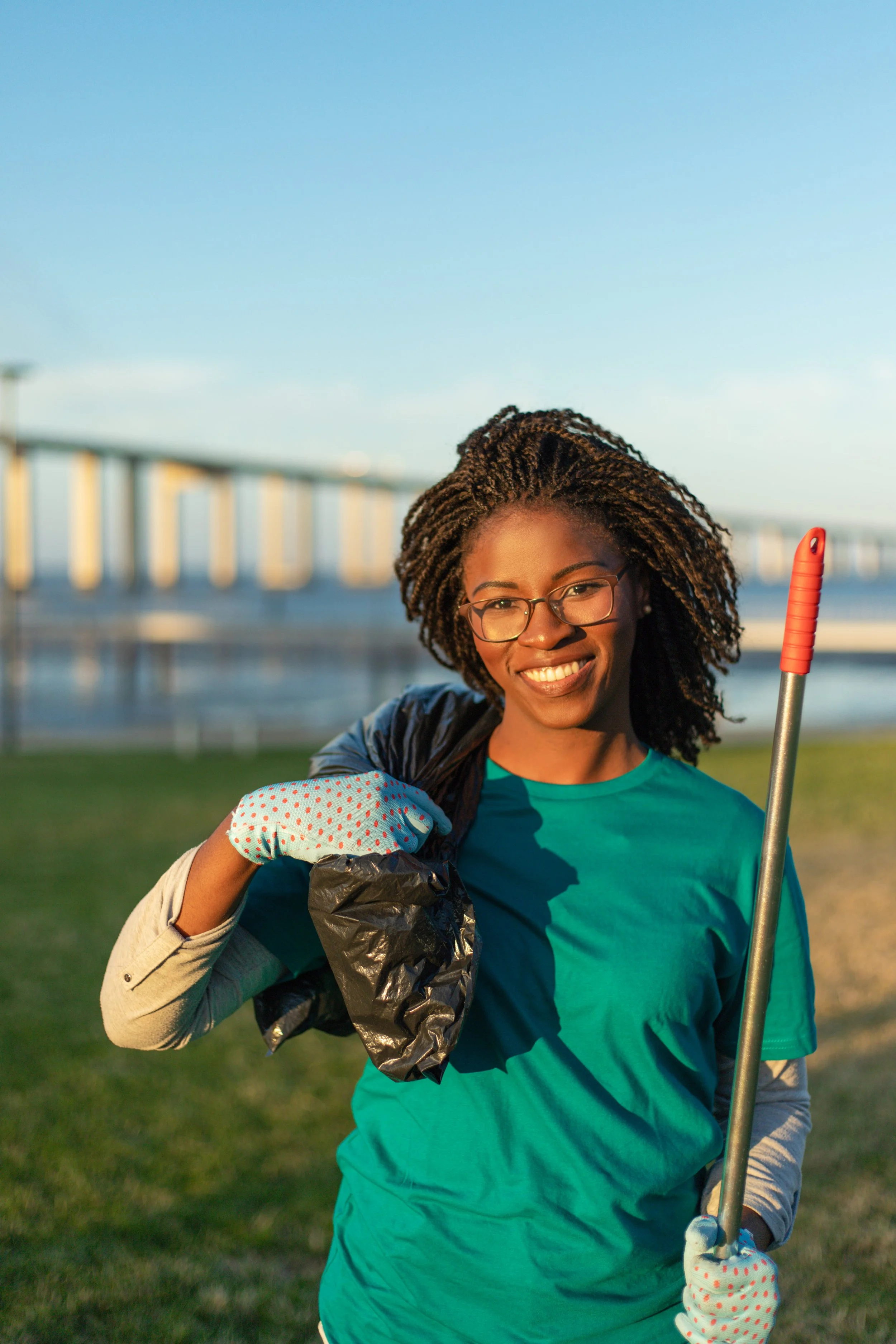 Smiling woman in a teal shirt wearing cleaning gloves, holding trash bag and litter picker outdoors near a bridge at sunset.