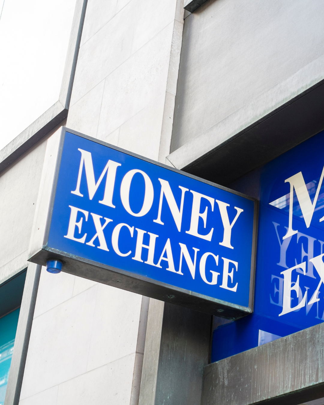 Blue sign with white text that reads "MONEY EXCHANGE" outside a building, with the sign's reflection visible on a glass surface.