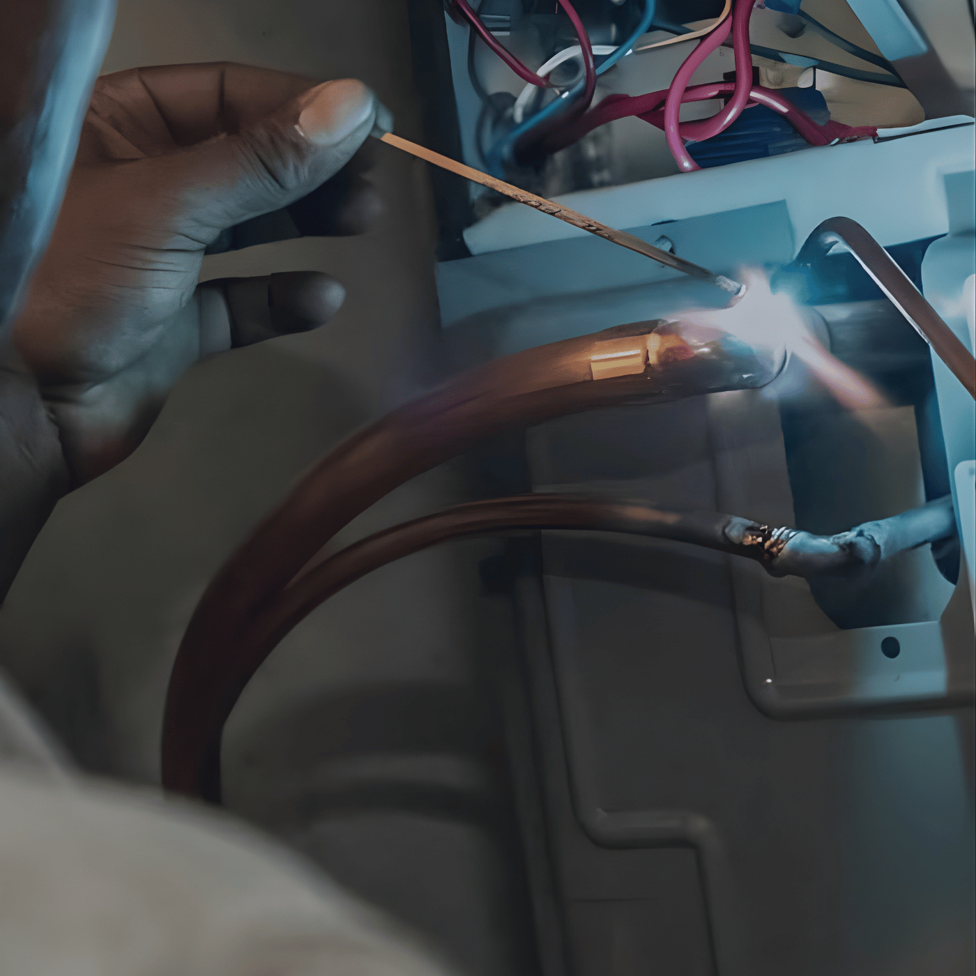 Close-up of a person's hand welding copper pipes inside an electrical panel, with wires and components visible in the background.