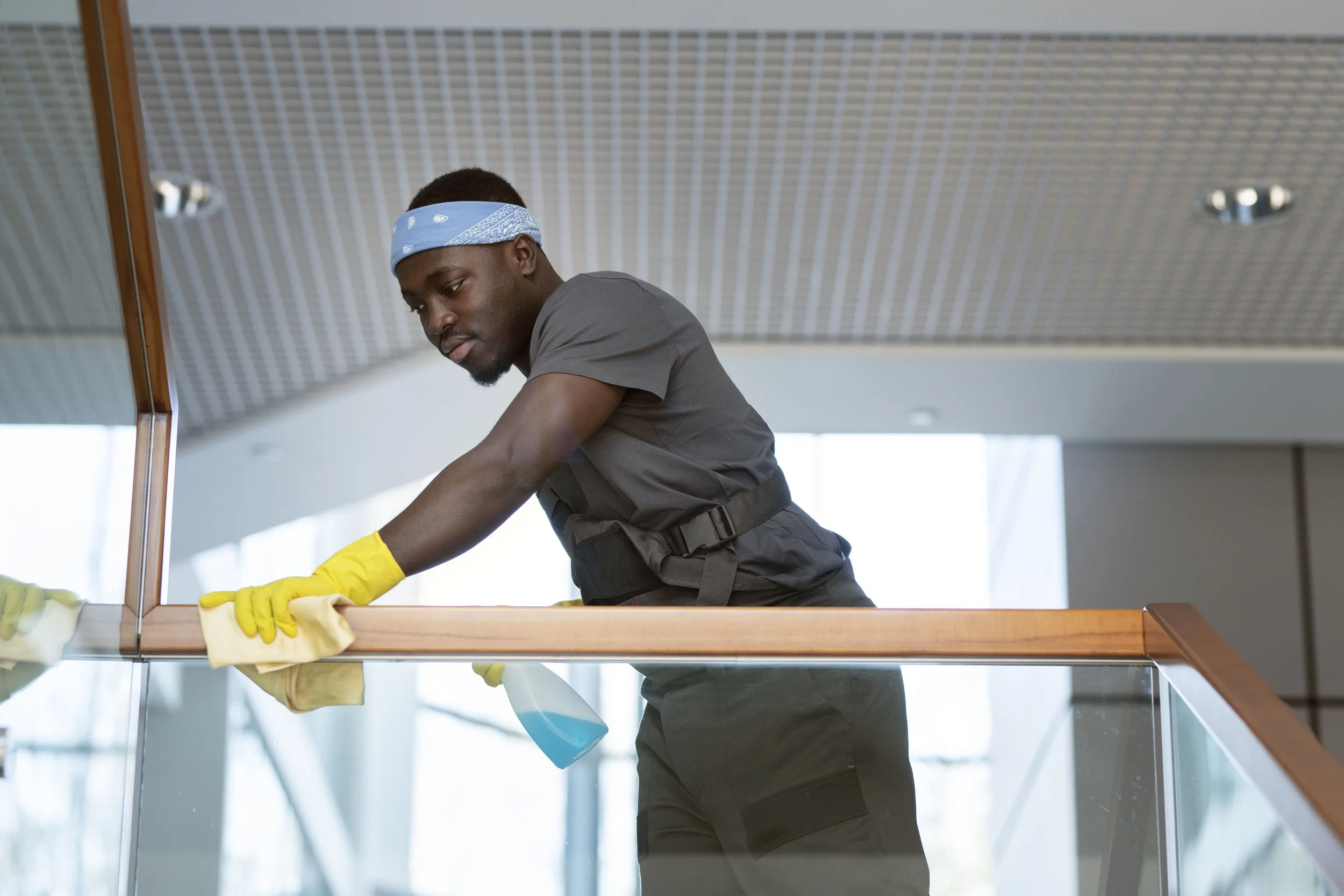 A man wearing a gray shirt, black pants, a blue bandana, and yellow cleaning gloves is cleaning a glass railing with a cloth and cleaning spray inside a modern building with large windows.