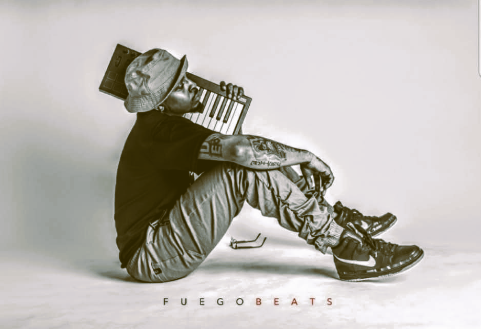 A person wearing a bucket hat, sunglasses, and casual clothing, sitting on the floor with legs bent, holding a small keyboard or piano. The person's tattoos are visible on their arm, and they are wearing Nike sneakers. The background is plain and light-colored, with