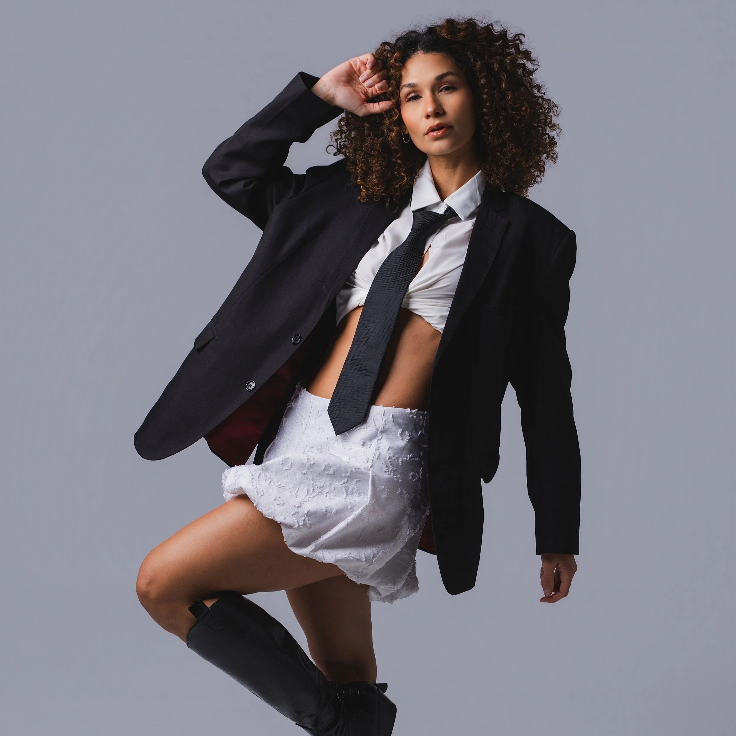 Fashion model wearing a black blazer, white crop top, black tie, white lace skirt, black knee-high boots, posing against a gray background.