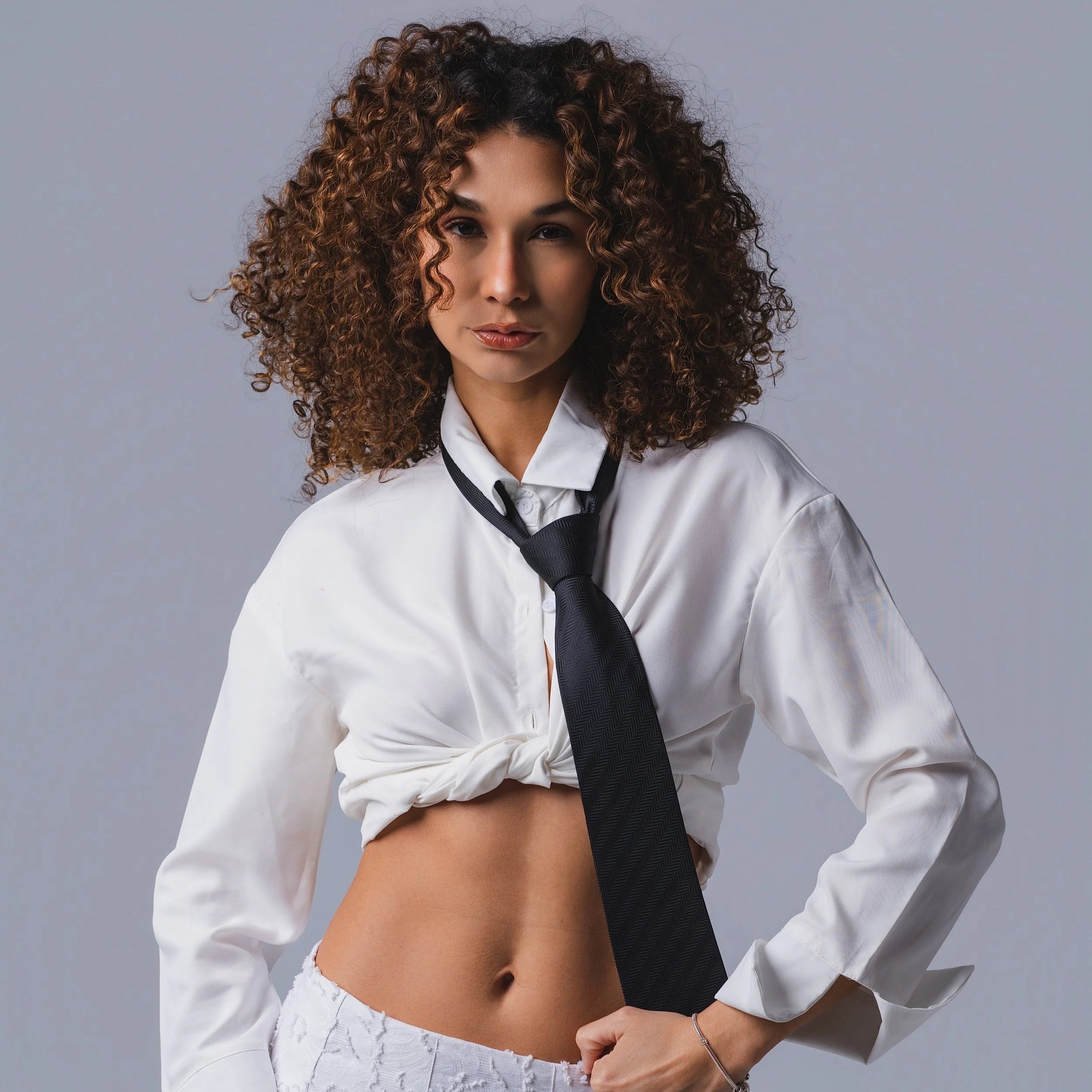 A woman with curly brown hair, wearing a white shirt tied at the waist, a black necktie, and white shorts, posing against a plain grey background.