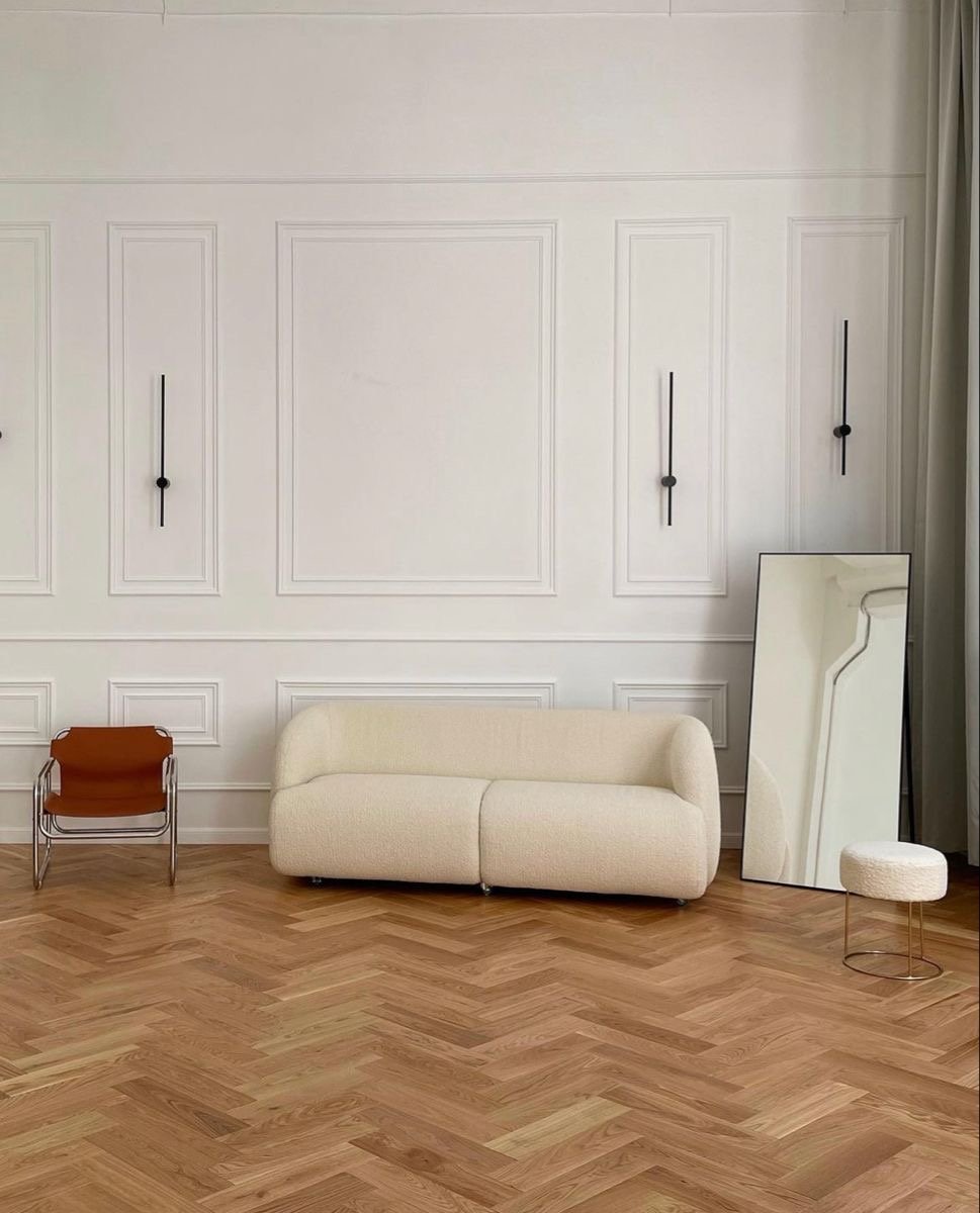 Minimalist interior with a white sofa, a large mirror leaning against the wall, a small round ottoman, a brown chair, and wall-mounted black reading lights on a white paneled wall.