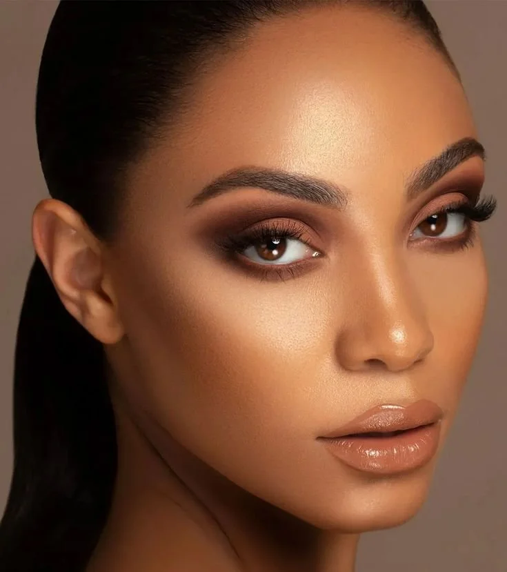 Close-up portrait of a woman with flawless skin, dark eye makeup, and nude glossy lips, looking at the camera.