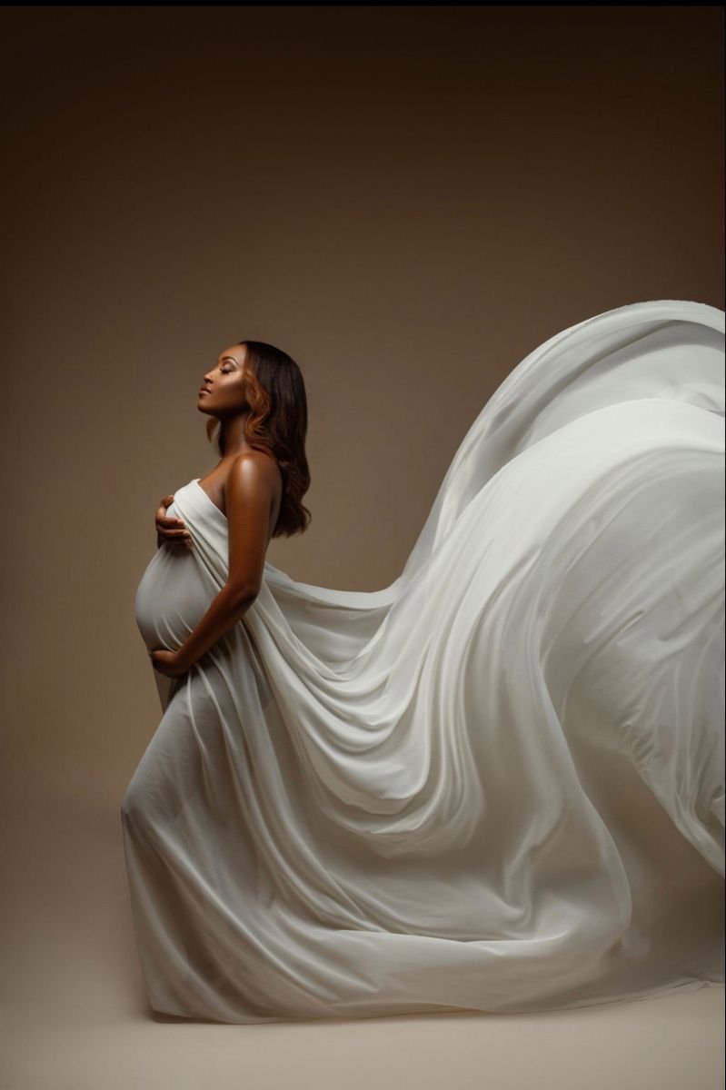 A pregnant woman posing elegantly wrapped in white fabric with flowing drapery in a studio setting.