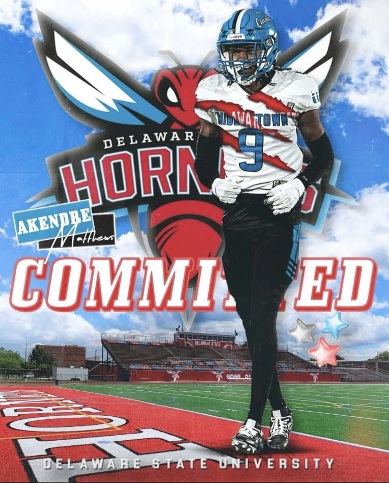 A football player in a Delaware State University uniform, wearing a blue helmet with a yellow stripe, standing on a football field with a red track. The background features a large Delaware Hornets logo with a red hornet, blue and white wings, and cl