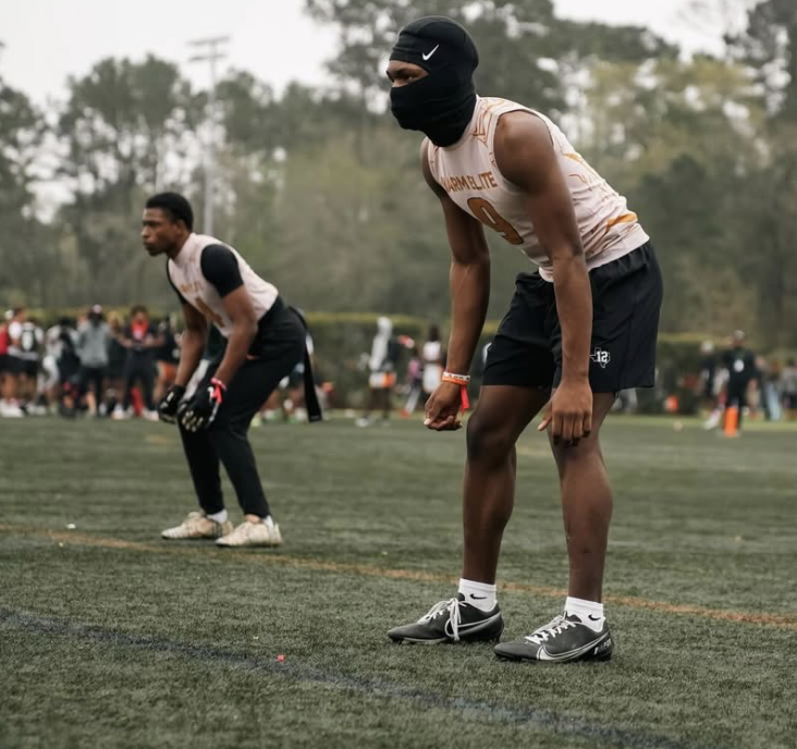 Two football players standing on the field, one in the foreground wearing a black mask and black shorts, and one in the background in a similar outfit, preparing for a play during a game.