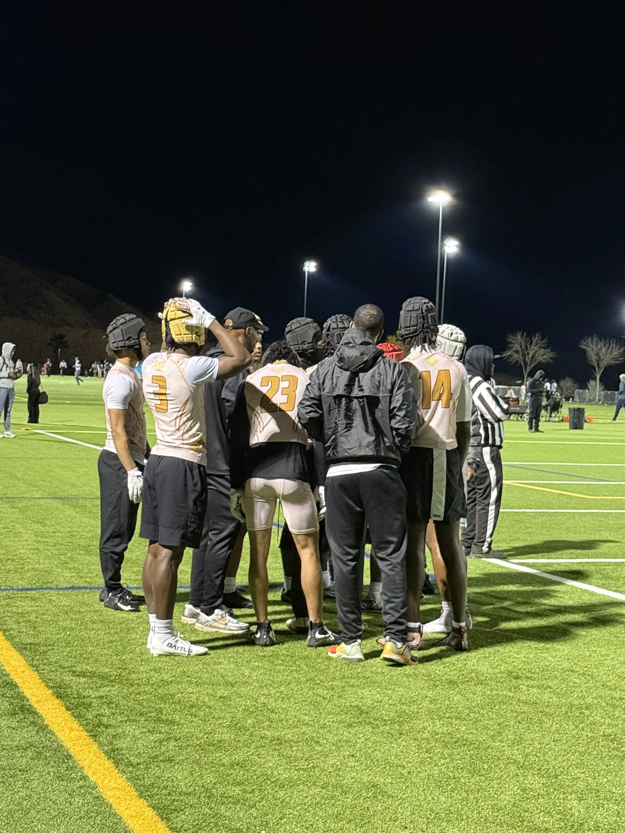 Football players in jerseys and helmets gather on a lit-up field at night, likely during a game or practice.