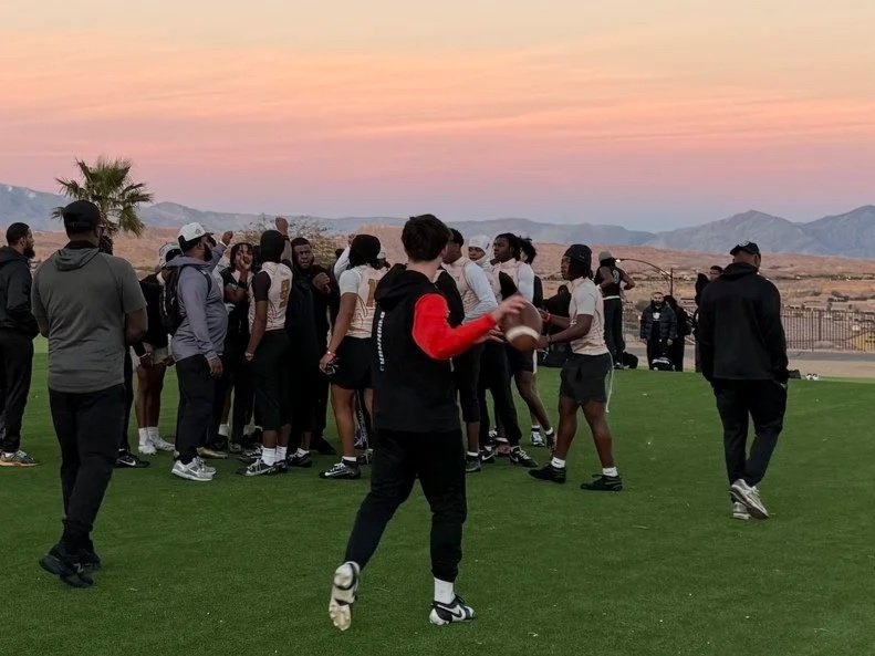 Group of young athletes on a grassy field during sunset, some holding footballs, with mountains in the background.