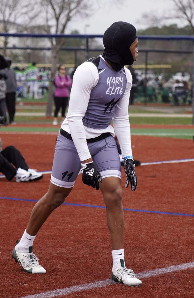 A female athlete wearing a black head covering, athletic clothing with the words 'HARM ELITE' on her chest, and gloves, stands on a running track during a sports event with other spectators and athletes in the background.