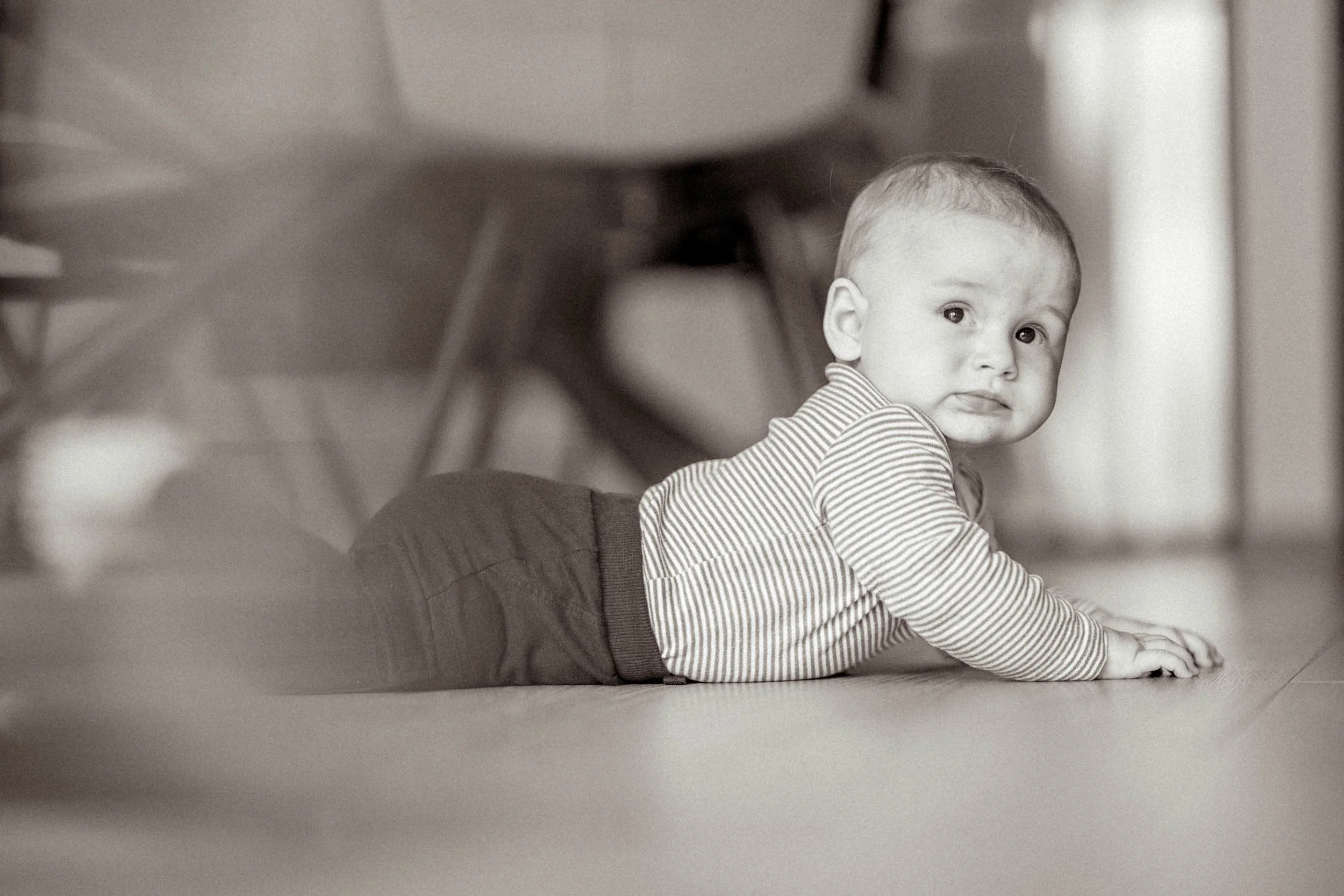 Ein Baby liegt auf dem Bauch auf einem Boden und schaut zur Kamera, im Hintergrund Möbel und ein Stuhl, Schwarz-Weiß-Fotografie.