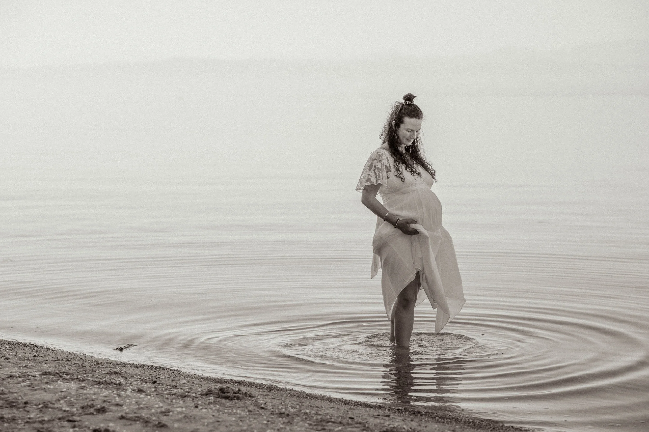 Schwangere Frau in einem weißen Kleid, die im Wasser an einem Strand steht und auf ihren Bauch schaut.