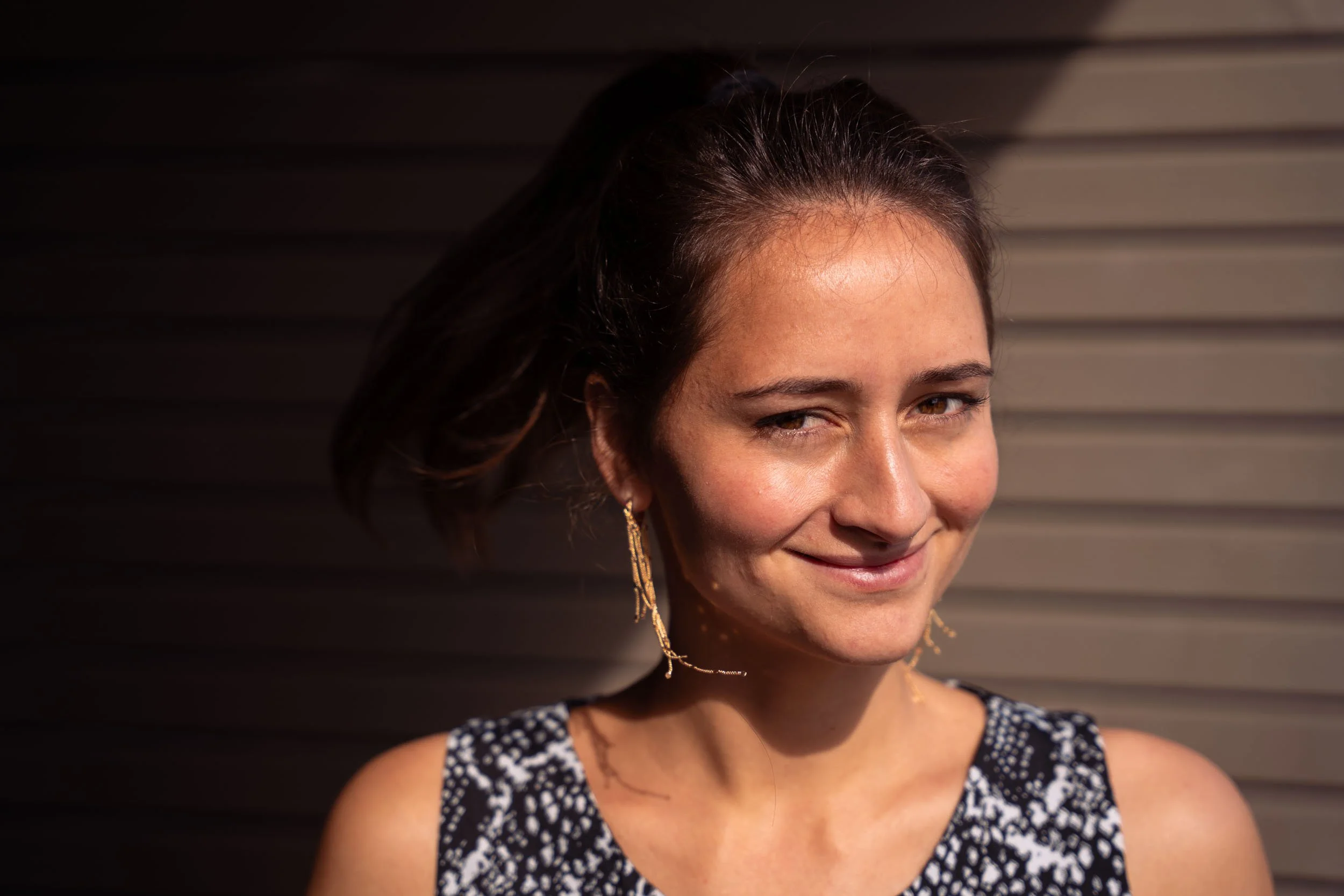 Eine junge Frau mit braunen Haaren und langen Ohrringen, die nach rechts schaut, vor einem dunklen, gestreiften Hintergrund.
