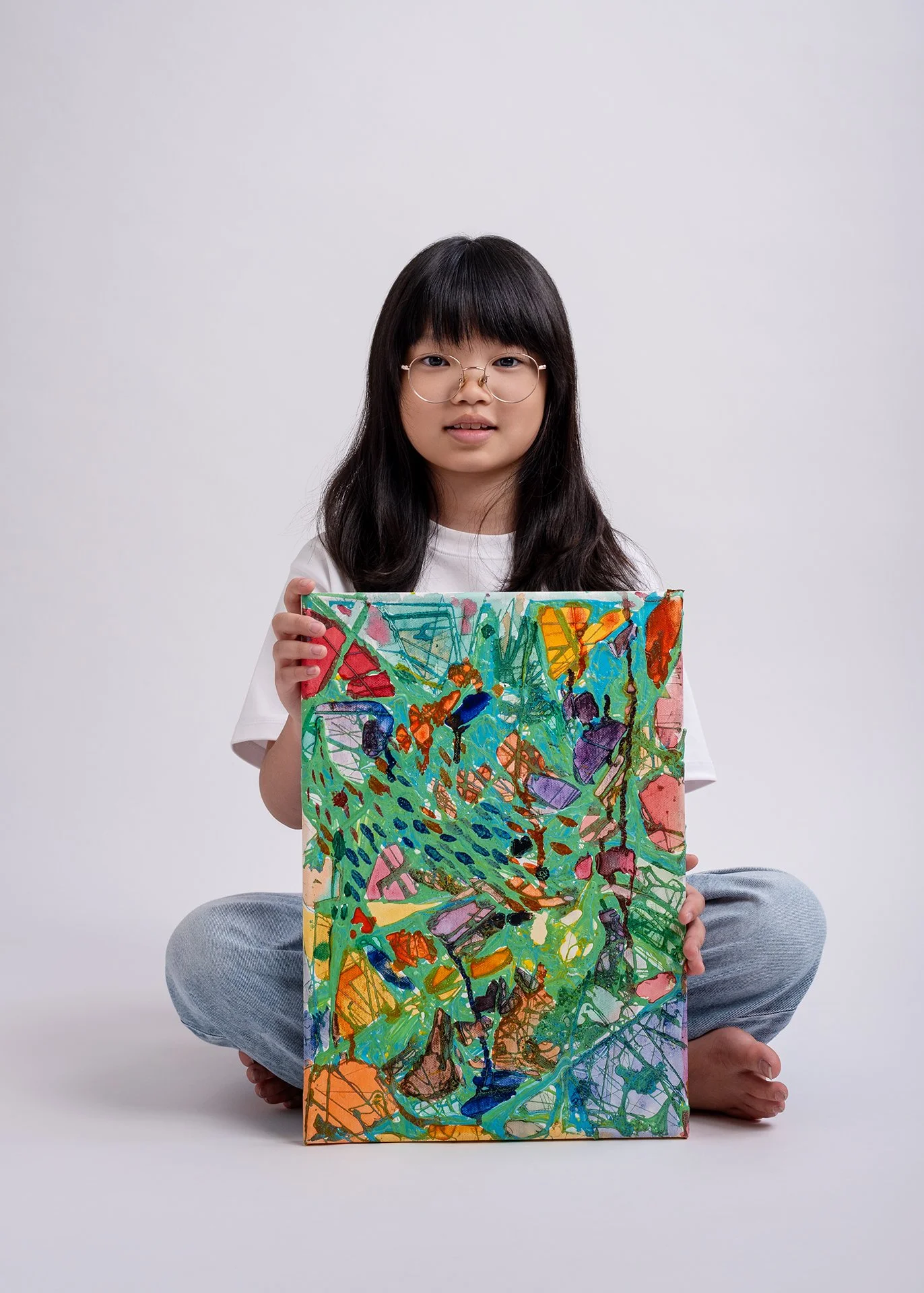 A young girl with glasses and long dark hair sits cross-legged on the floor, holding a colorful abstract painting in front of her. The background is plain white.