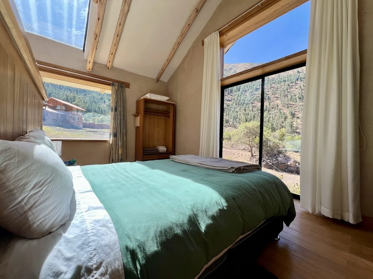 A cozy bedroom with a large bed featuring white pillows and a green bedspread, large windows showing a mountain landscape, light-colored curtains, and wood accents in the ceiling and furniture.