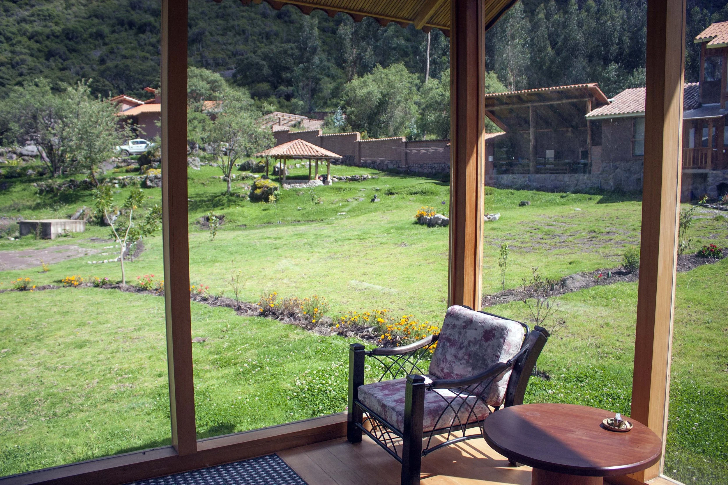 View of a lush green yard with small trees, flower beds, and a stone pathway, seen through a glass window from a porch with a chair and a small round table.