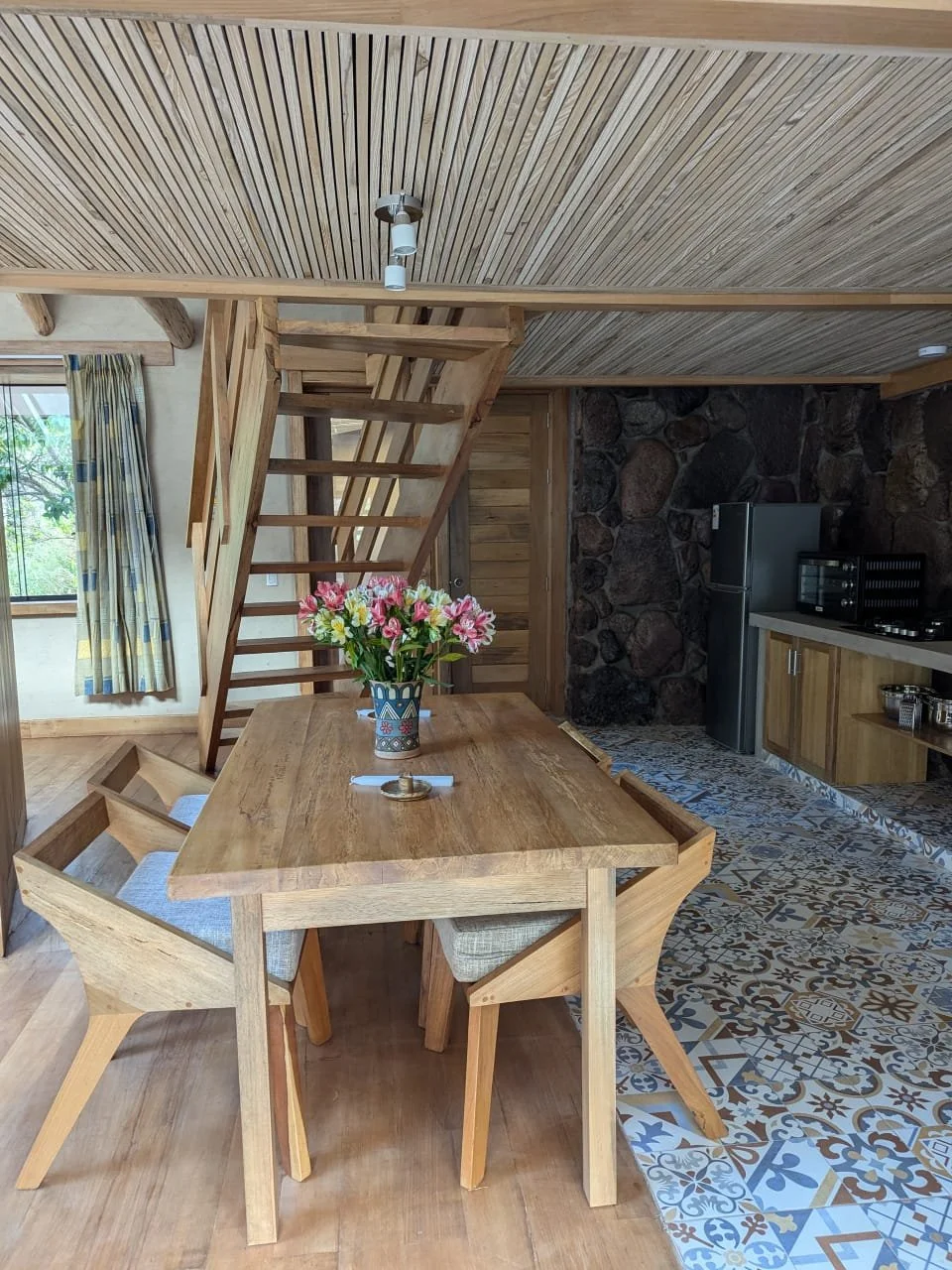 Wooden dining table with a vase of pink and white flowers, four chairs with cushioned seats, in a rustic room with wooden ceiling, staircase, and a stone wall kitchen area with a refrigerator and patterned tile floor.