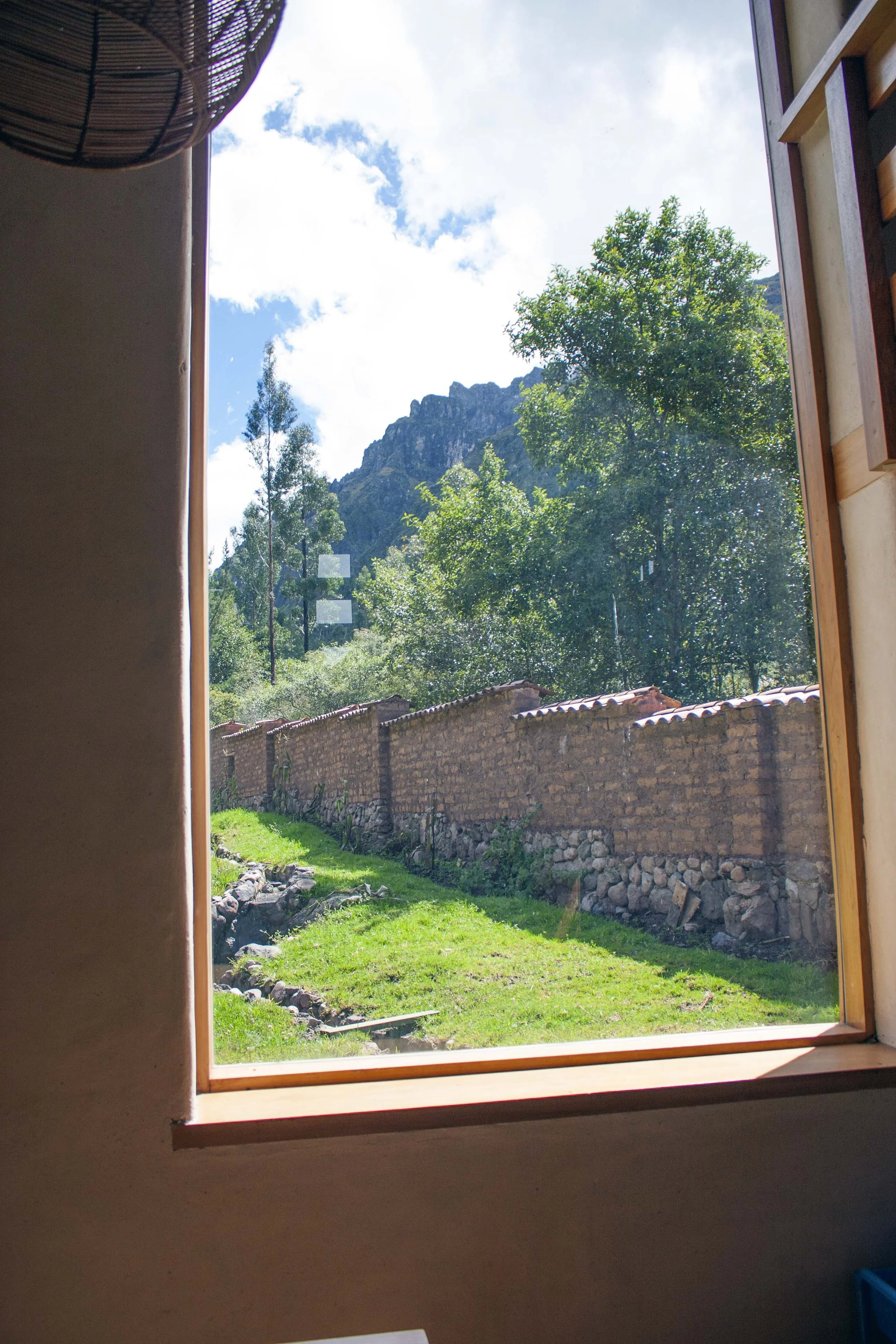 View through a window showing a grassy yard with a stone and brick wall, trees, and a mountain in the background under a partly cloudy sky.