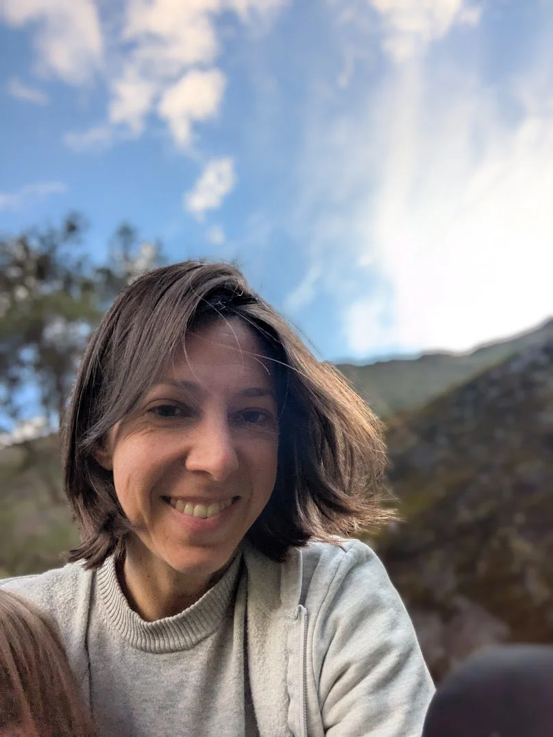 A woman smiling outdoors with a blue sky and clouds in the background.