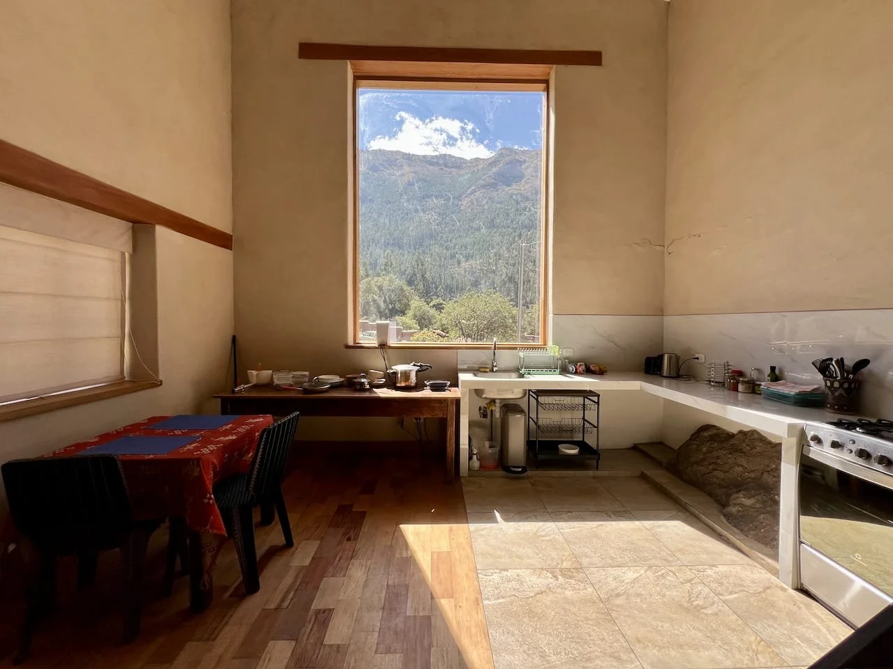 A kitchen with a large window showing a mountain landscape outside, with a table covered with a red tablecloth, chairs, and kitchen appliances along the countertop.