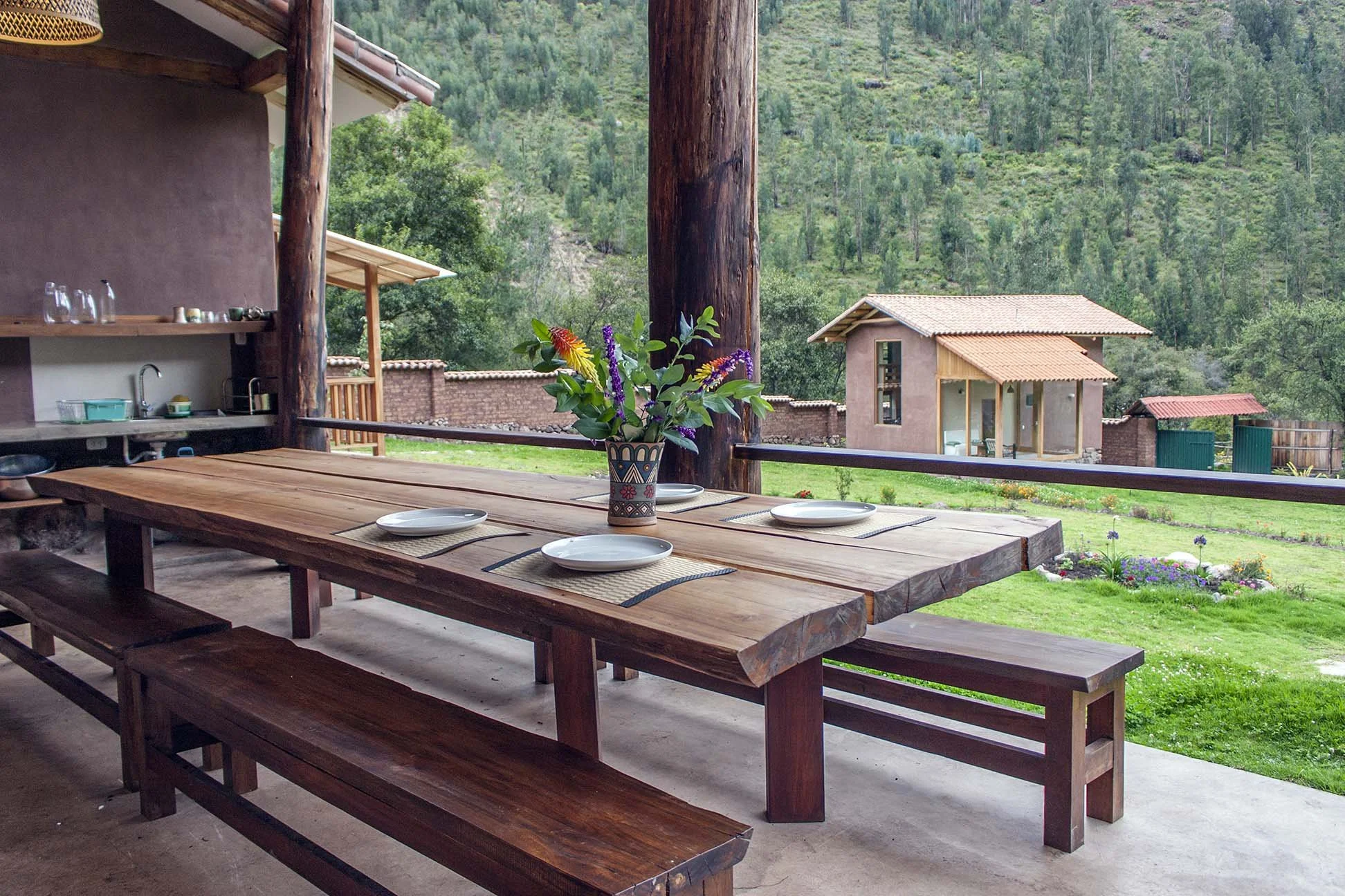 A rustic outdoor dining table set with four white plates and a colorful flower arrangement in a patterned vase on a covered porch overlooking a green landscape with hills and small buildings.