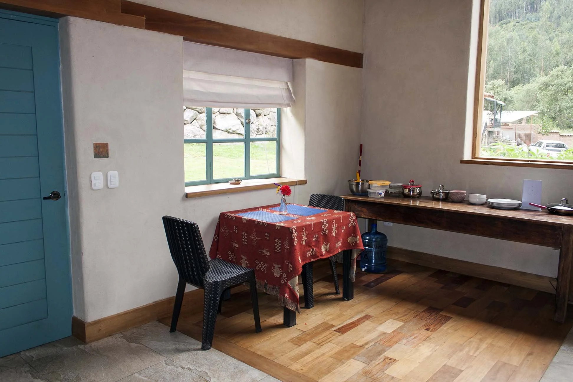 A cozy kitchen corner with a small dining table covered with a red patterned tablecloth, two black chairs, and a wooden counter with kitchen items. There are two large windows, one dressed with a white shade, overlooking a lush green outdoor area.