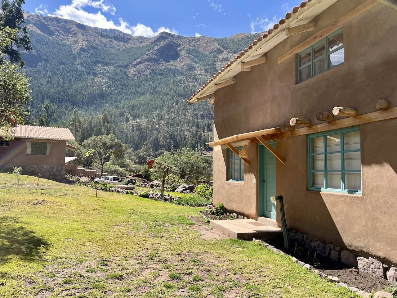 A rustic house with turquoise window frames and a green door, set against a backdrop of mountains and green trees, with a grassy yard in the foreground.