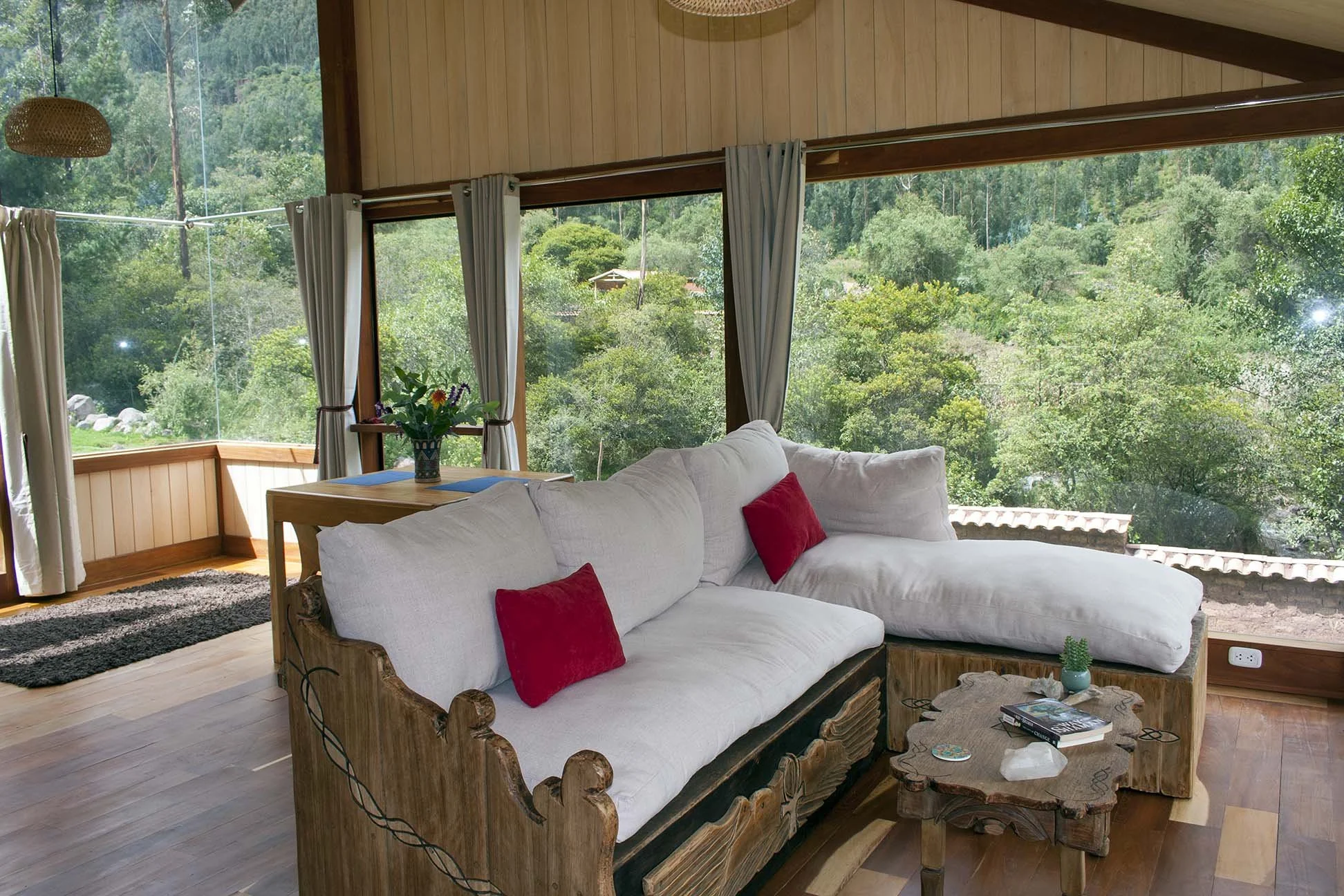 Living room with large windows showing a green forest outside, white sectional sofa with red pillows, small wooden coffee table with books and decor, and a dining table with flowers.