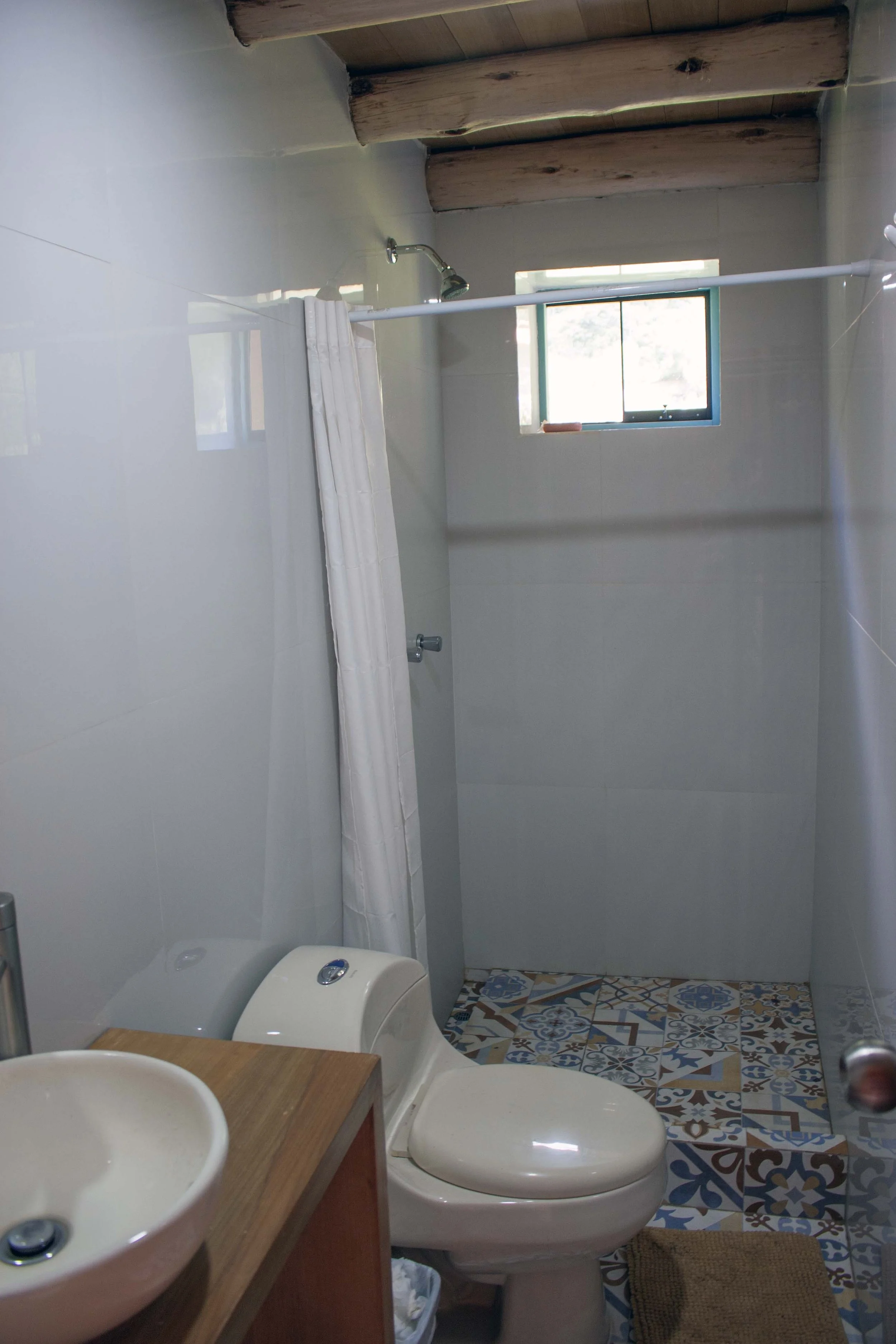 A small bathroom with a toilet, a wooden sink, and a white shower curtain. The shower area has a small window and a showerhead. The floor has colorful patterned tiles. The ceiling has exposed wooden beams.