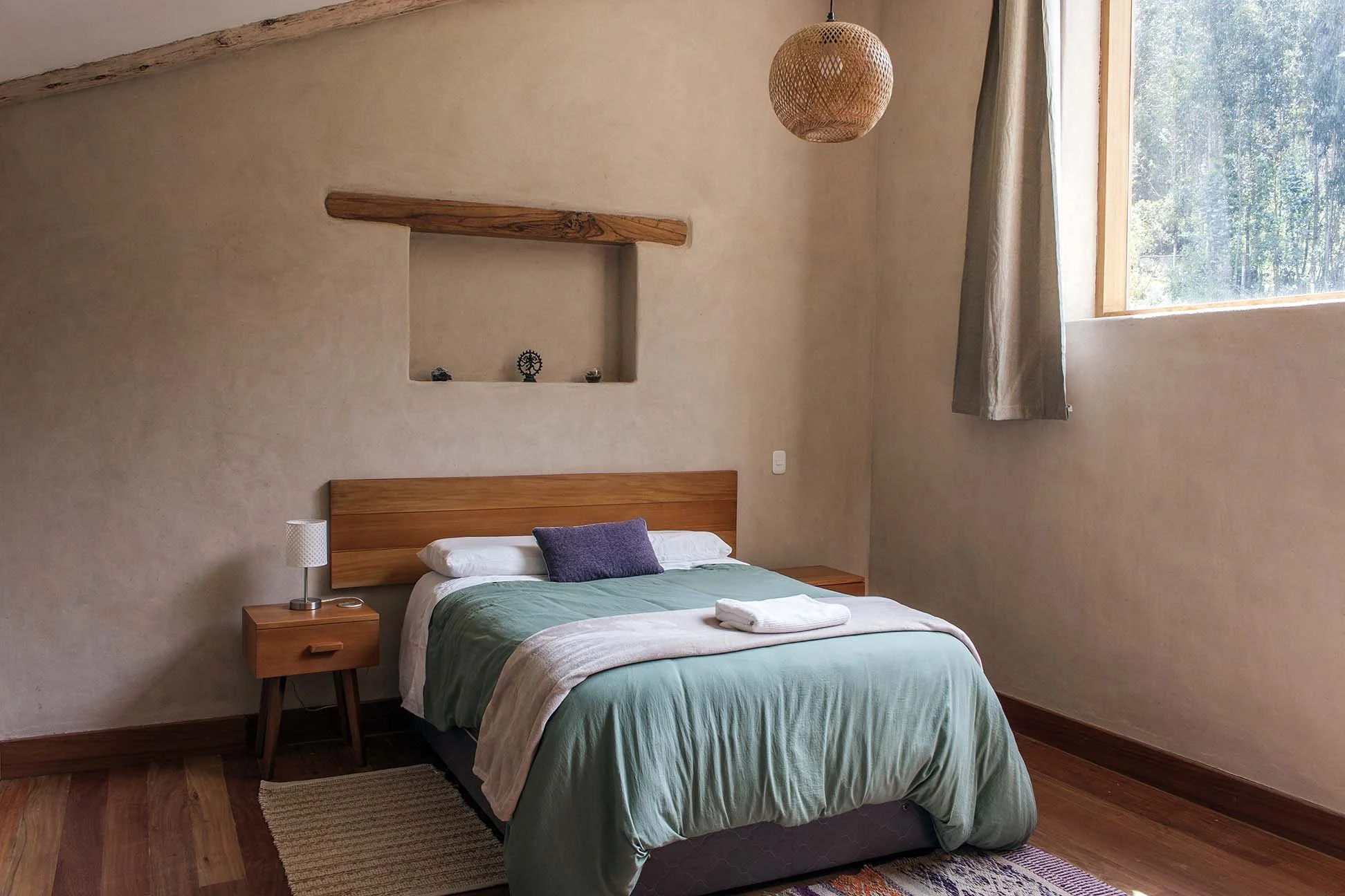 A bedroom with a wooden bed, green bedding, and a purple pillow. A small wooden nightstand with a lamp is next to the bed. The wall behind the bed has a built-in wall niche with decorative objects. A large window with beige curtains lets in natural light, and a woven pendant light hangs from the ceiling.