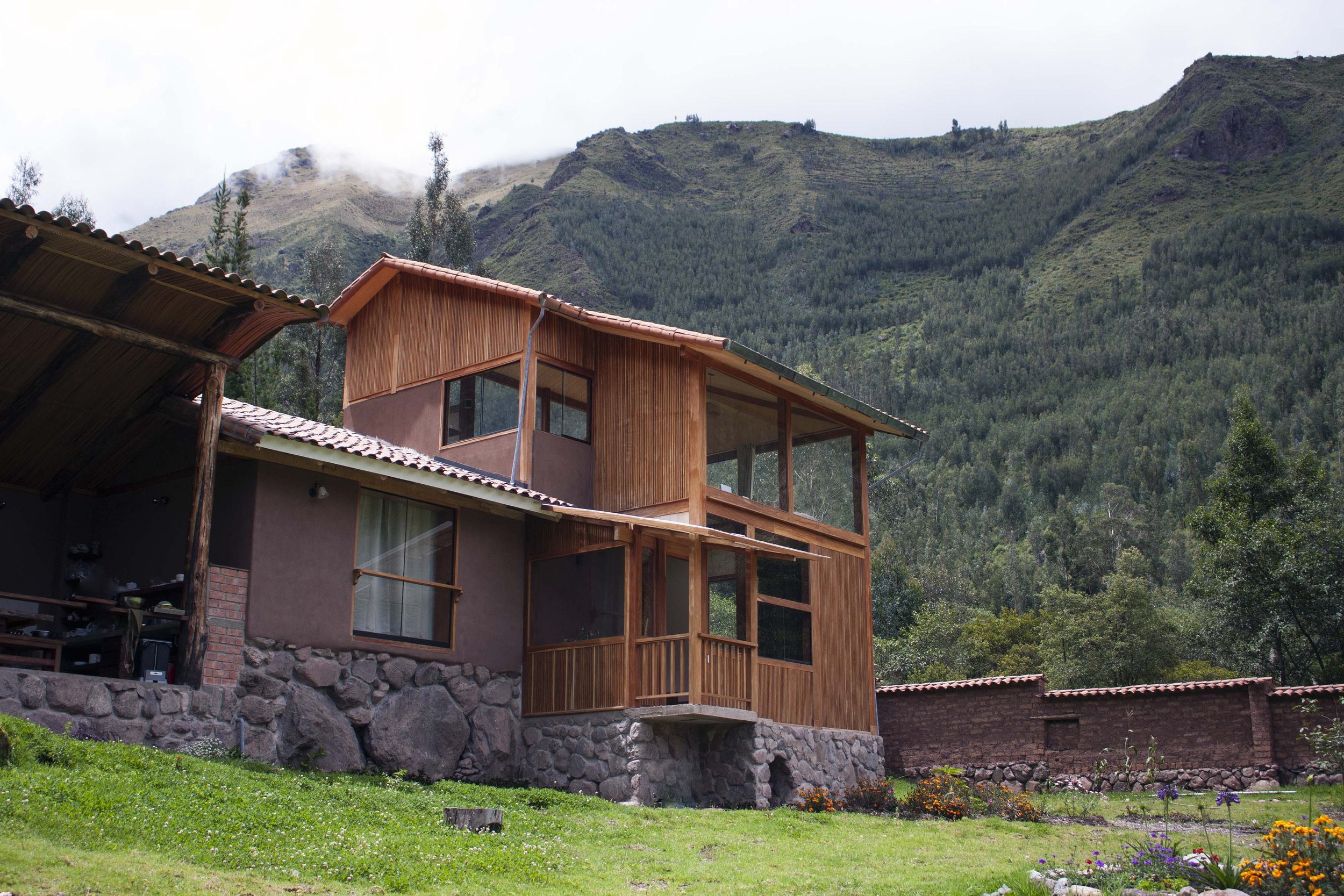 A modern house made of wood and stone, situated on a grassy lawn with colorful flowers, against a backdrop of lush green mountains and cloudy sky.