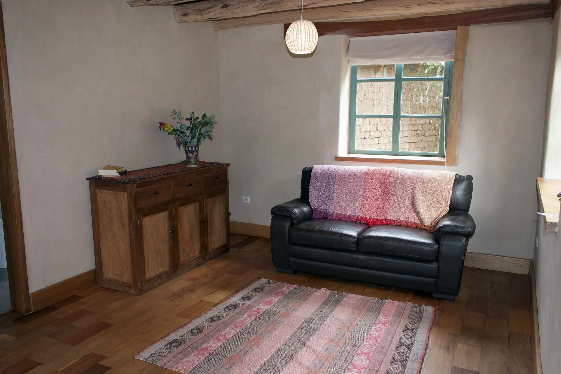 A cozy living room with a black leather sofa covered by a pink and white gradient throw, a wooden sideboard with a vase of flowers, a colorful rug, a window with a view of brick wall outside, and a ceiling light