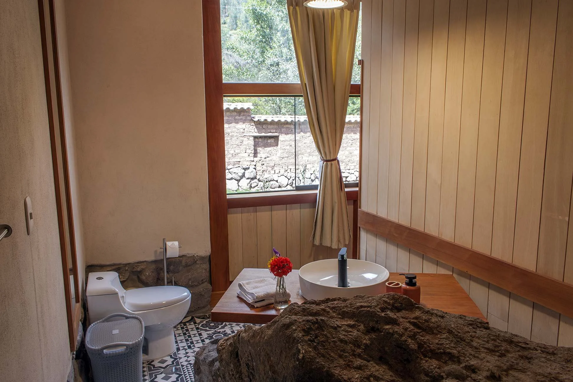 Rustic bathroom with a wooden vanity, a round white vessel sink, a beige curtain tied back, and a large rock in the foreground, with a window showing trees outside.