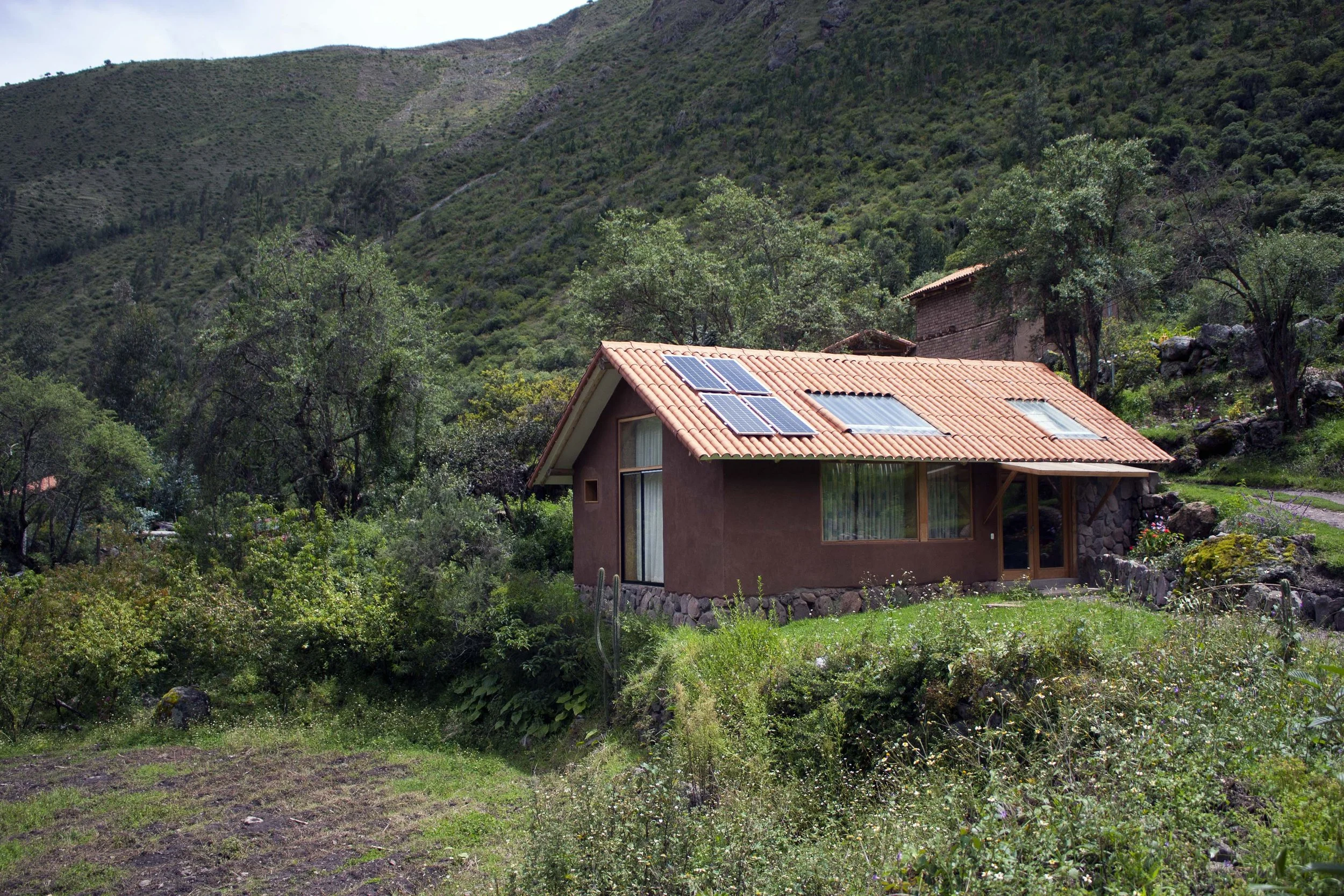 A house with solar panels on its red-tiled roof, located in a lush, green hillside with trees and rocky terrain in the background.