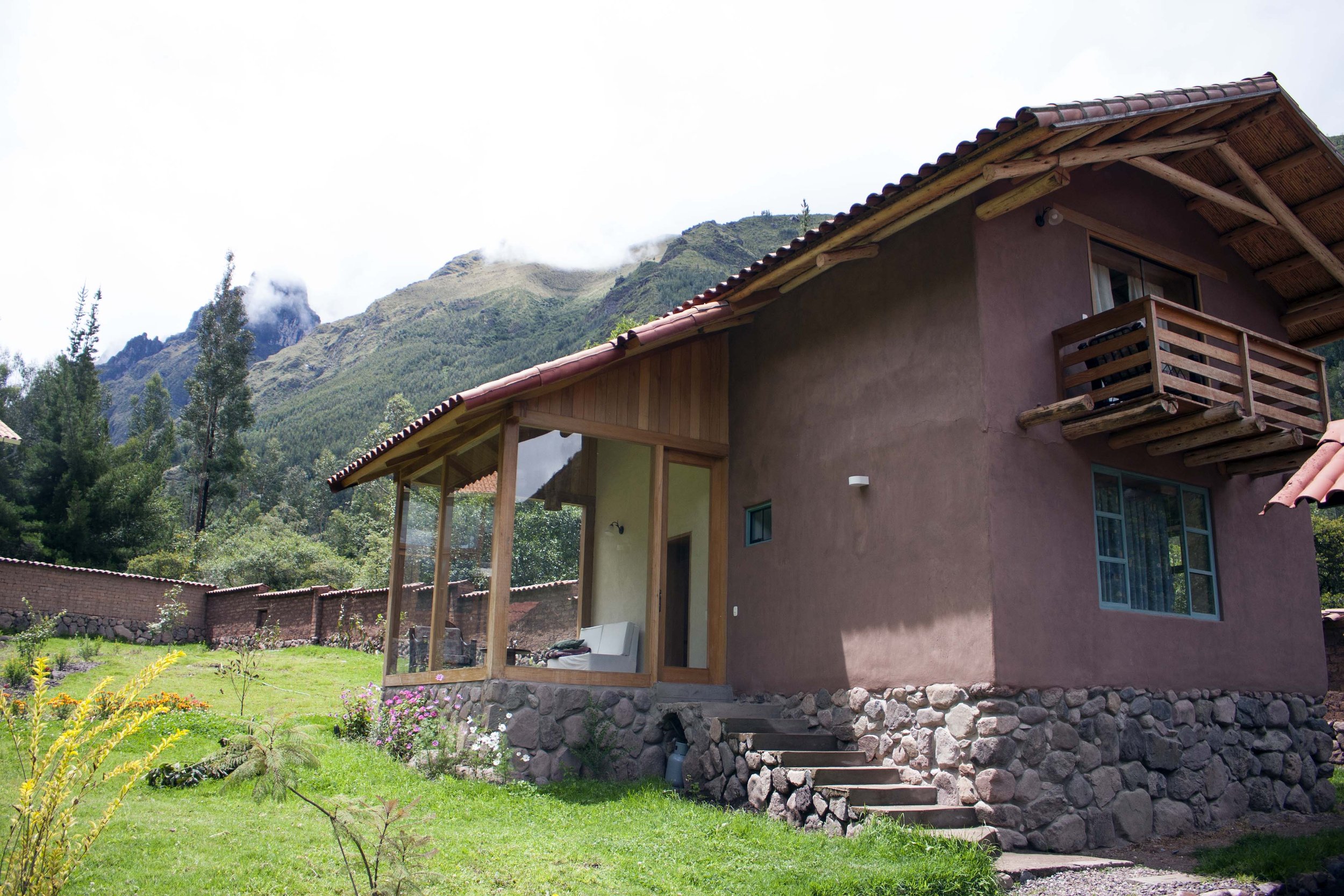 A two-story house with a stone foundation, stucco walls, and a tiled roof, featuring a balcony, surrounded by green grass and mountain scenery in the background.