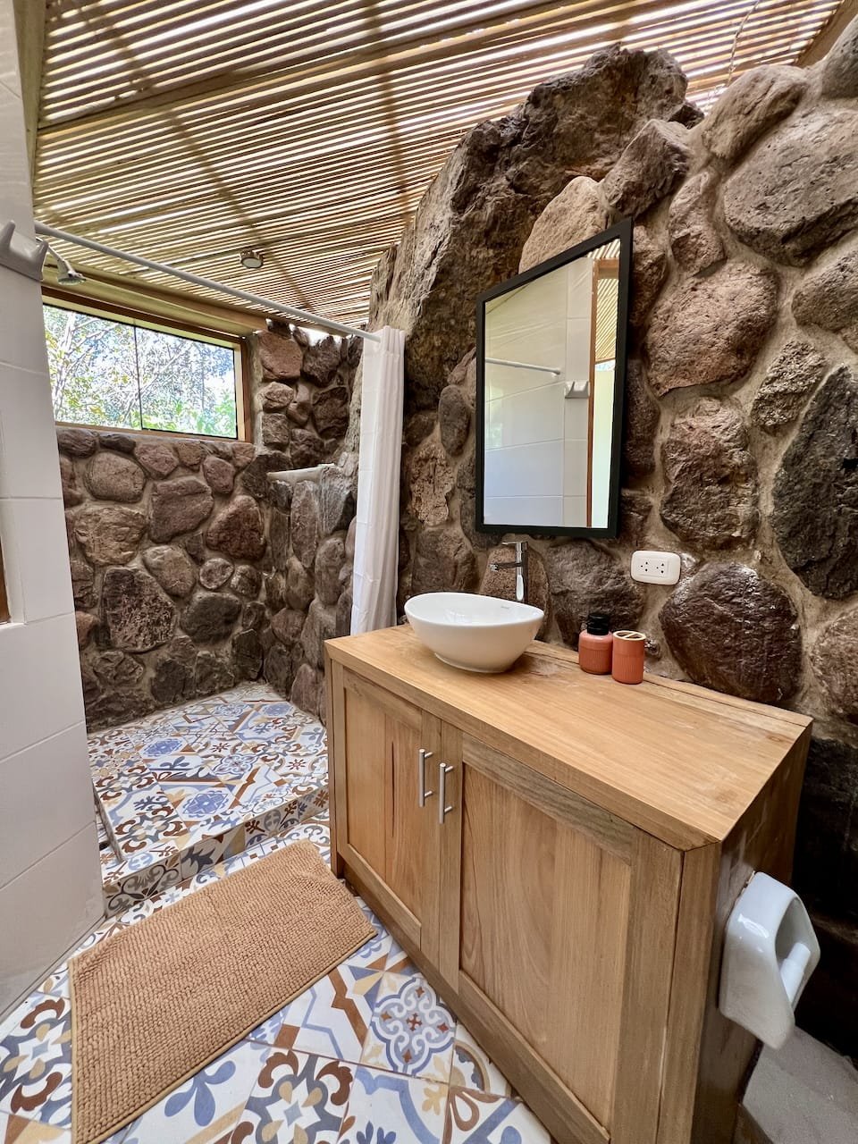 Rustic bathroom with stone walls, patterned tile floor, wooden vanity, vessel sink, mirror, and shower with curtain.