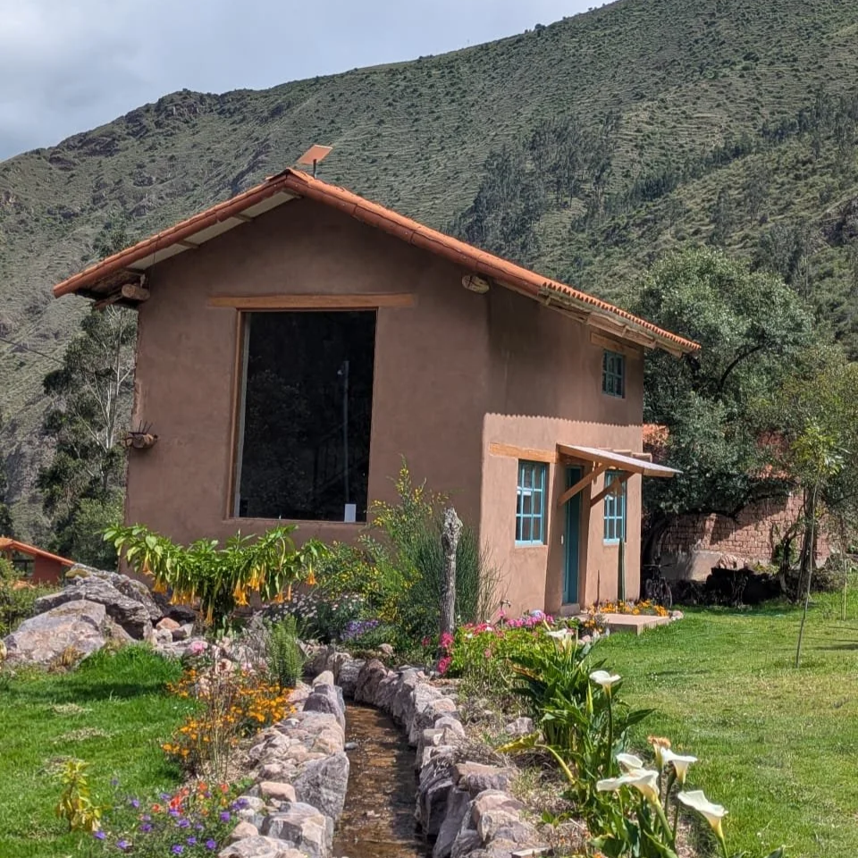 A rustic two-story house with a brown stucco exterior and a red tile roof, set against a lush mountain backdrop. The house has a large front window, a small porch with a wooden canopy, and blue window frames. A stone-lined garden path filled with colorful flowers surrounds the house.
