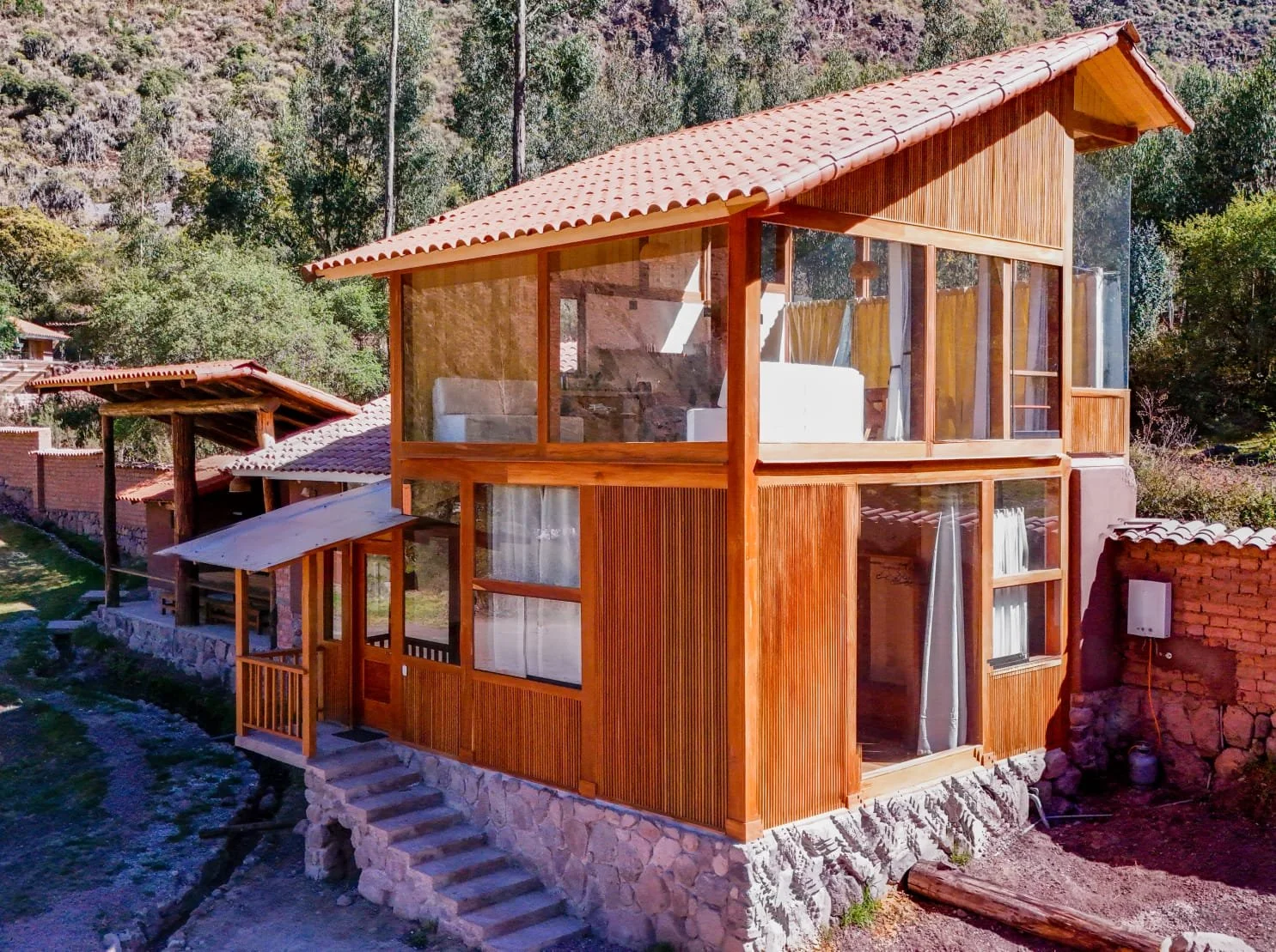 A two-story house with a wooden exterior and large glass windows, situated on a hillside with trees in the background.