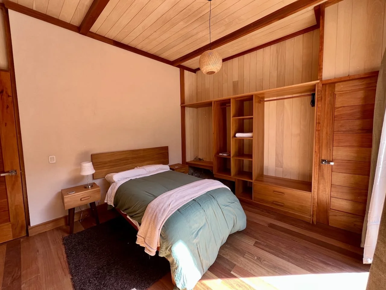 A bedroom with wooden walls, ceiling, and flooring, featuring a bed with green bedding, a small bedside table with a lamp, and an open wooden closet with shelves and drawers. Natural light coming in from a window on the right.
