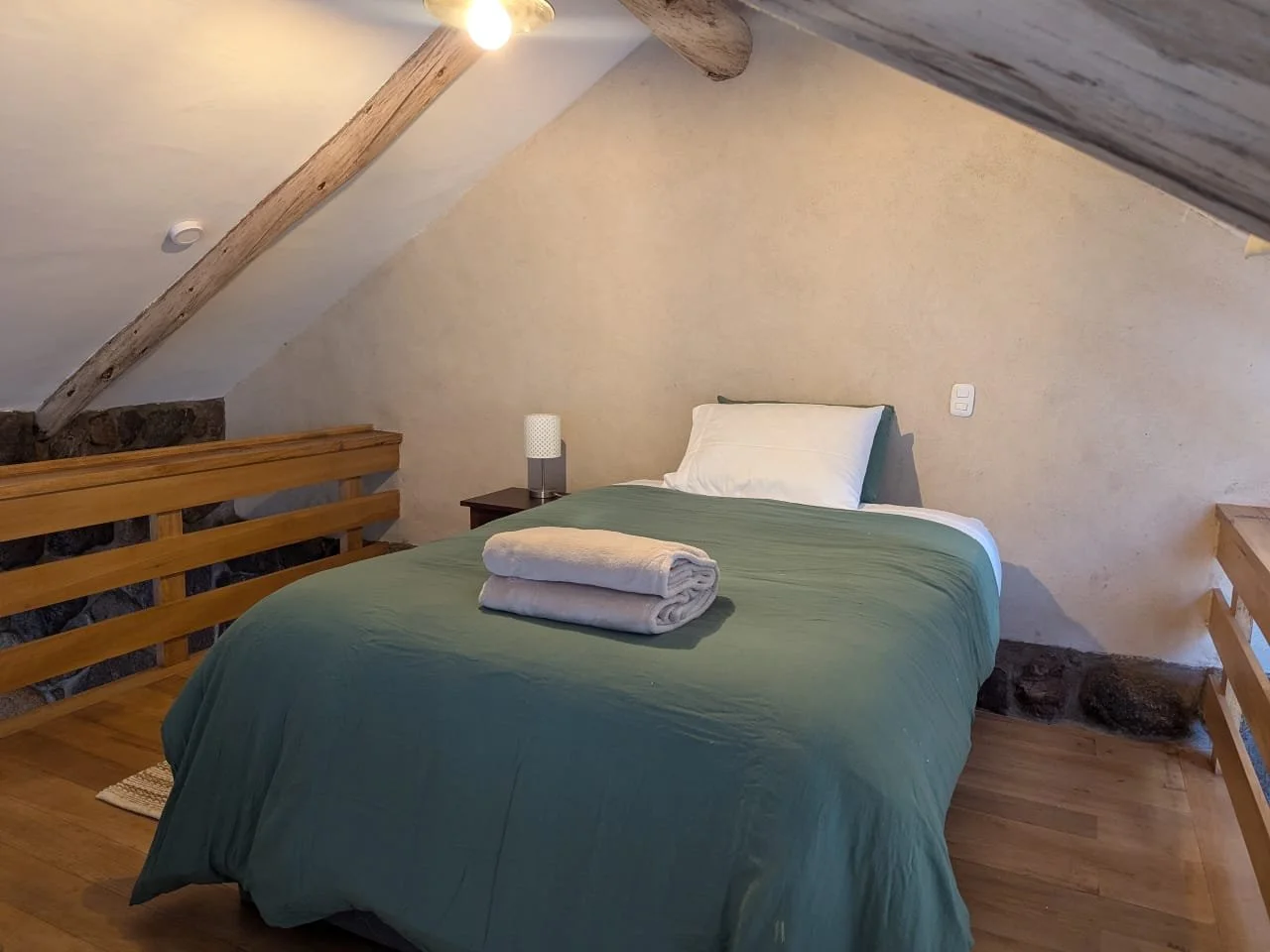 A cozy bedroom with a sloped ceiling, wooden beams, and a bed with green bedding, a white pillow, and folded towels on top. There's a nightstand with a lamp and a wooden railing along the side.
