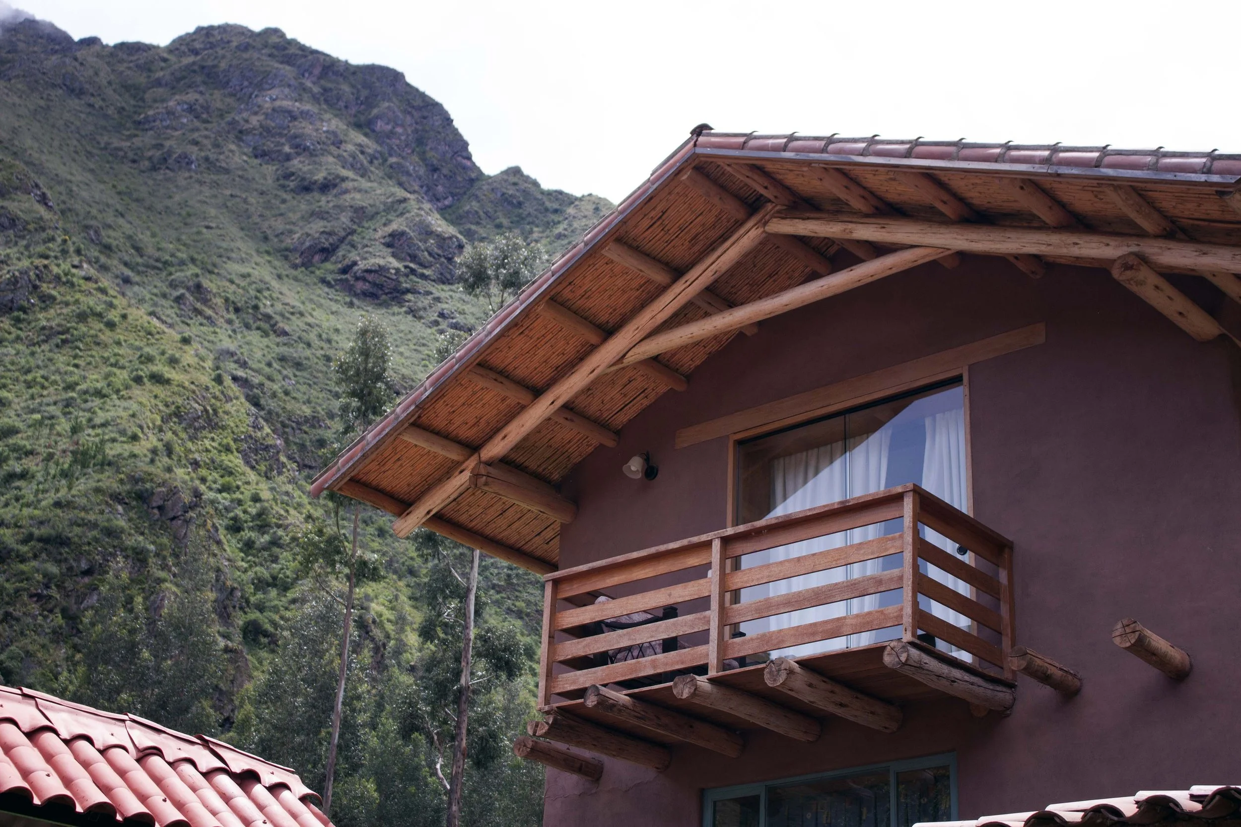 A house with a wooden balcony and a sloped roof, set against a green mountainous landscape.